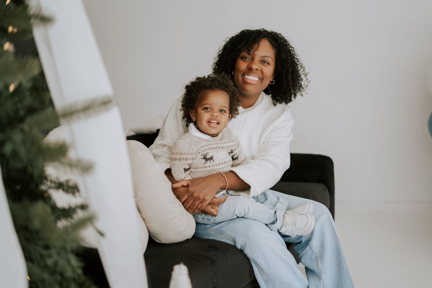 Smiling mother and toddler sit together on black chair beside a decorated Christmas tree.