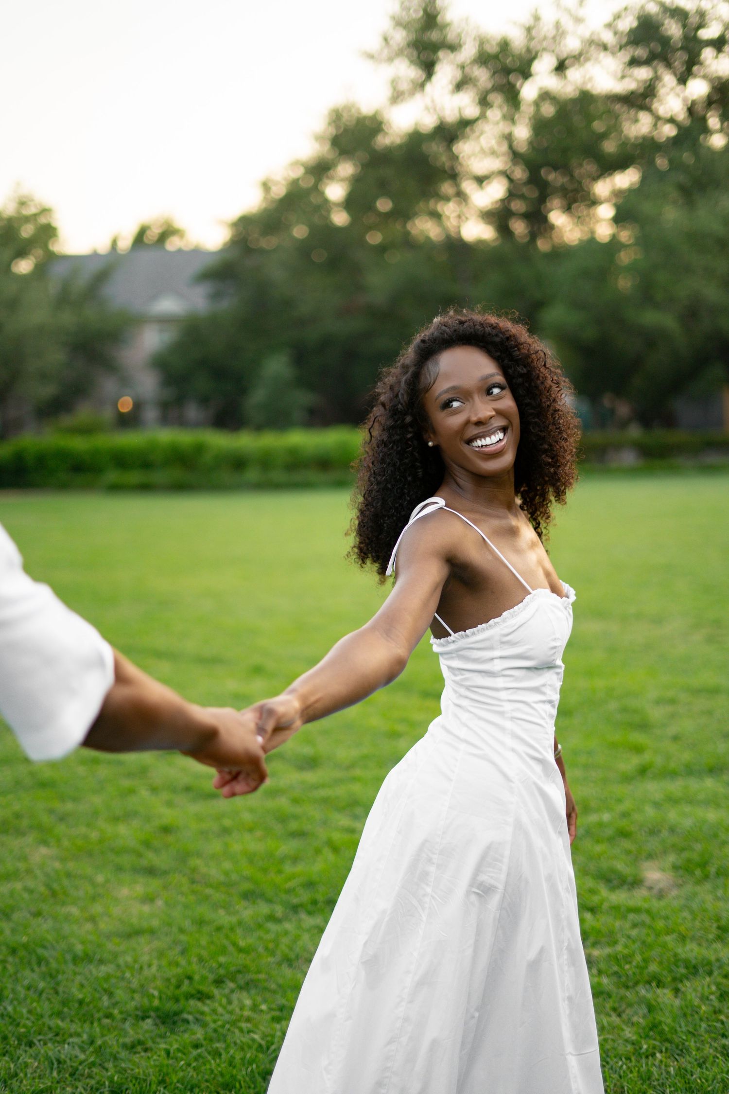 A beautiful Black woman in a white dress smiles radiantly while holding hands with her partner in a park.