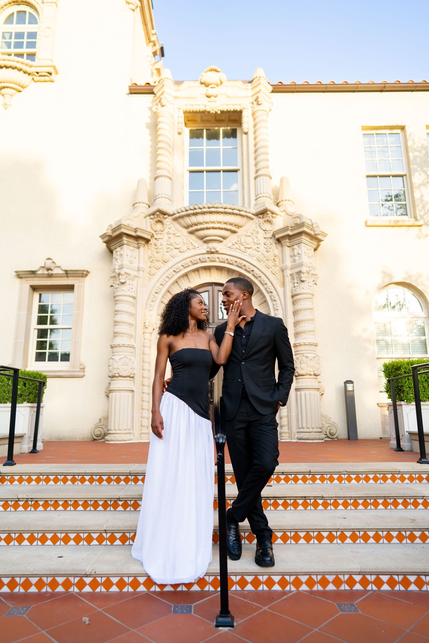 Elegant Black couple posing on ornate Spanish-style building steps, woman in black and white gown, man in black suit.