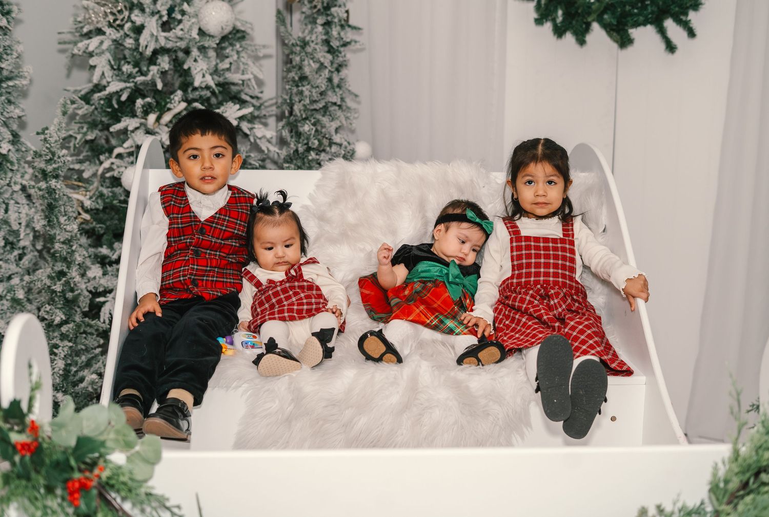 Four children dressed in red and green holiday outfits sit in a white sleigh with Christmas decor.
