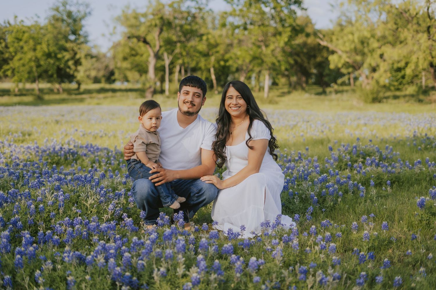 Family of three smiling together in a beautiful Texas bluebonnet field with green trees backdrop.