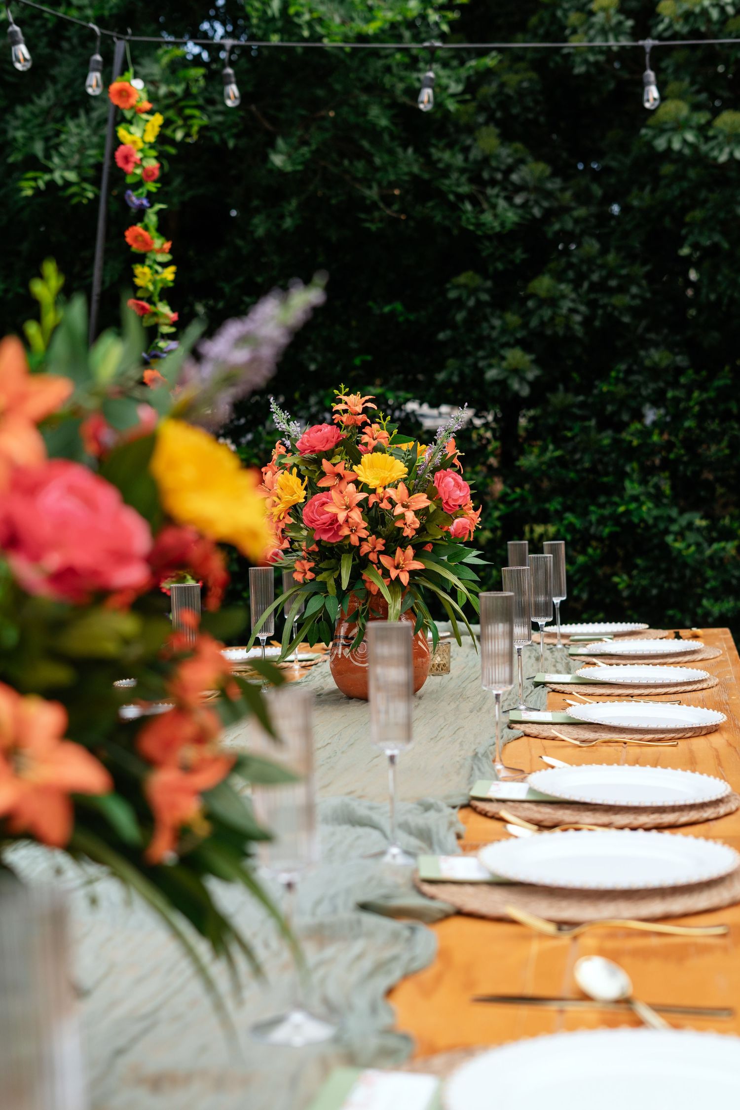 Outdoor dining table with colorful floral centerpieces, white plates, and string lights surrounded by lush greenery.