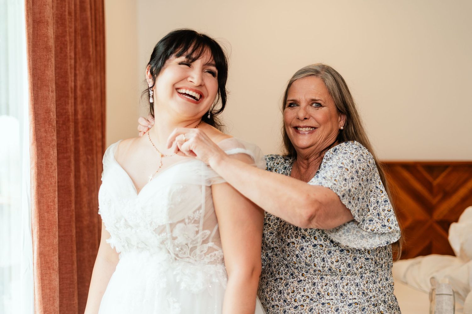 Bride laughing joyfully as older woman helps fasten her white lace wedding dress in a bright, elegant room.