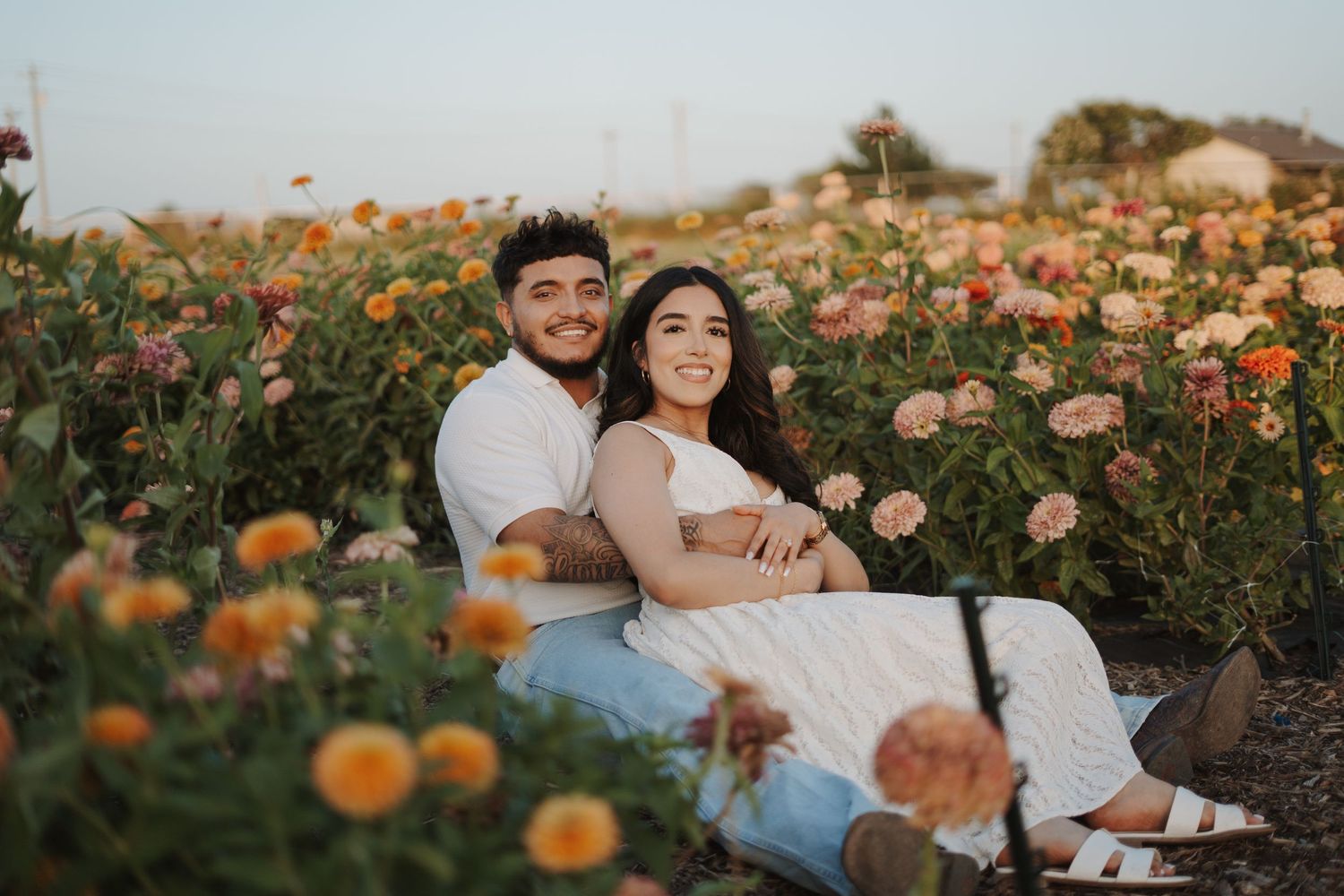 Couple sitting together in a colorful flower field, smiling, surrounded by pink and orange blooms at golden hour.