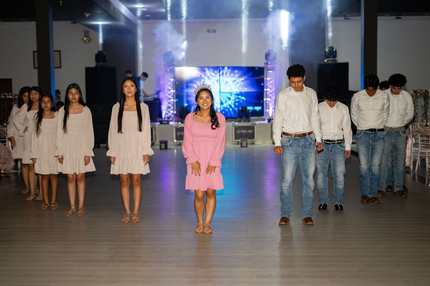 Quinceañera court of honor in white dresses and jeans lines up on dance floor before performance.