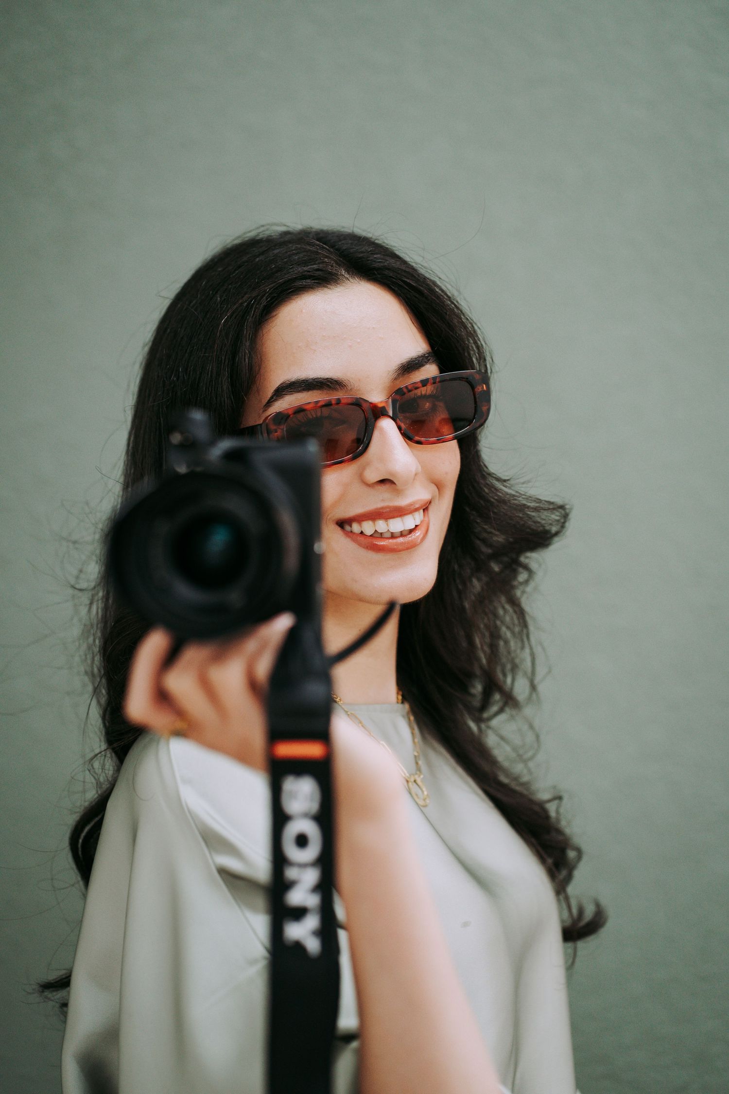 Smiling woman with dark hair and sunglasses holding a Sony camera against a light green wall.