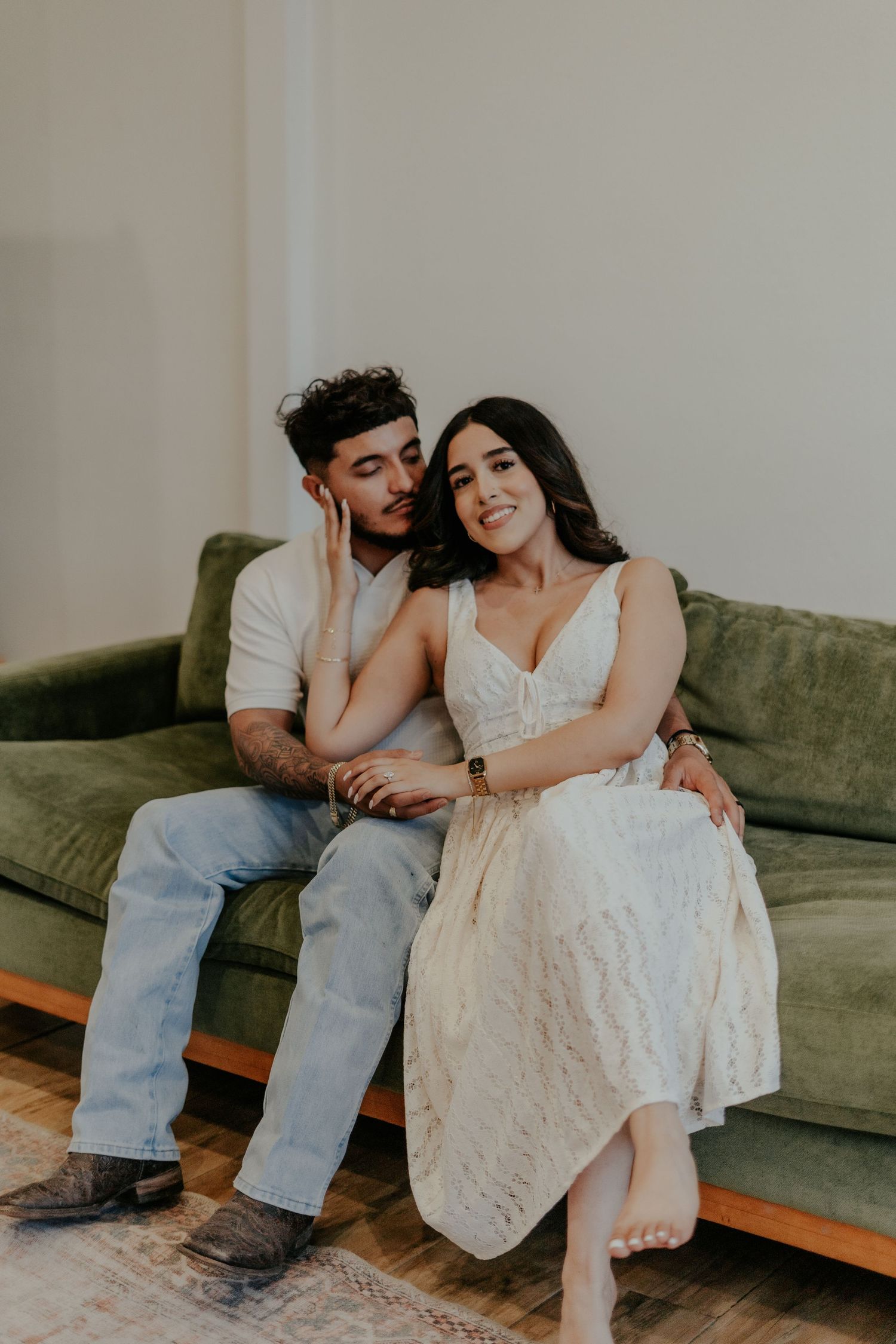 Couple sharing a tender moment on a green velvet couch, woman in white lace dress smiling softly.