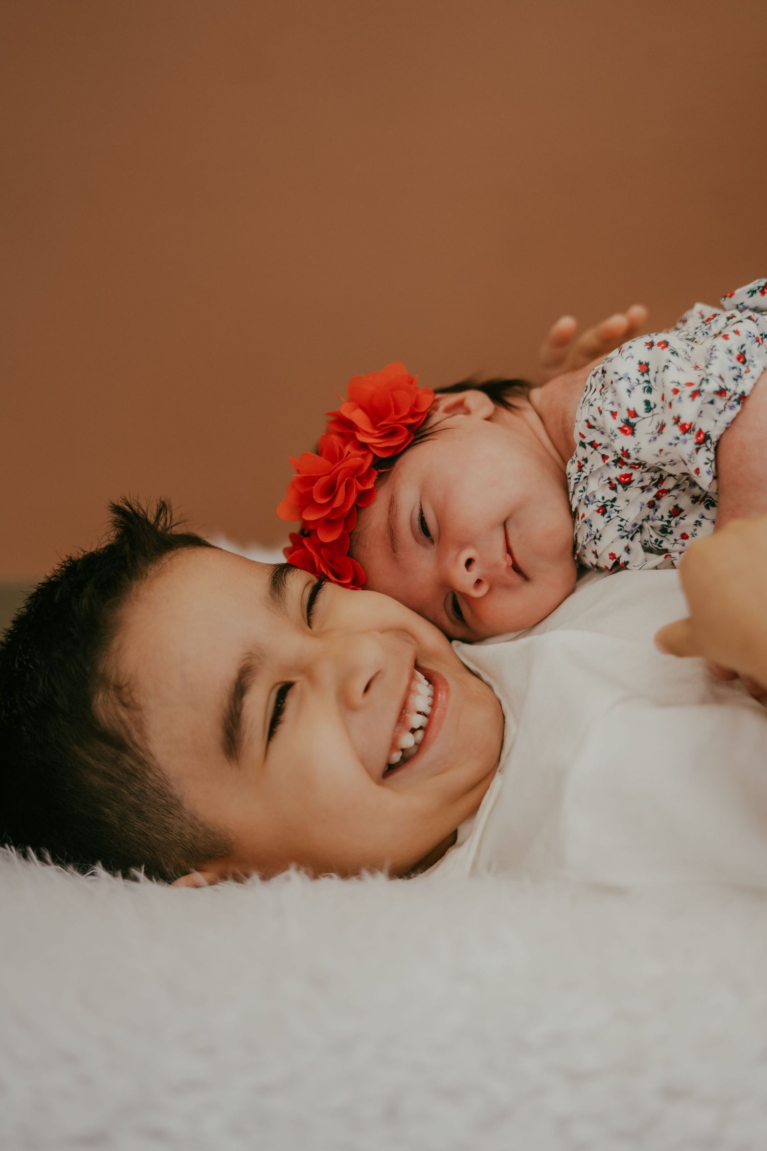 Smiling older sibling lies on white fur with newborn baby girl wearing red floral headband resting against their cheek.