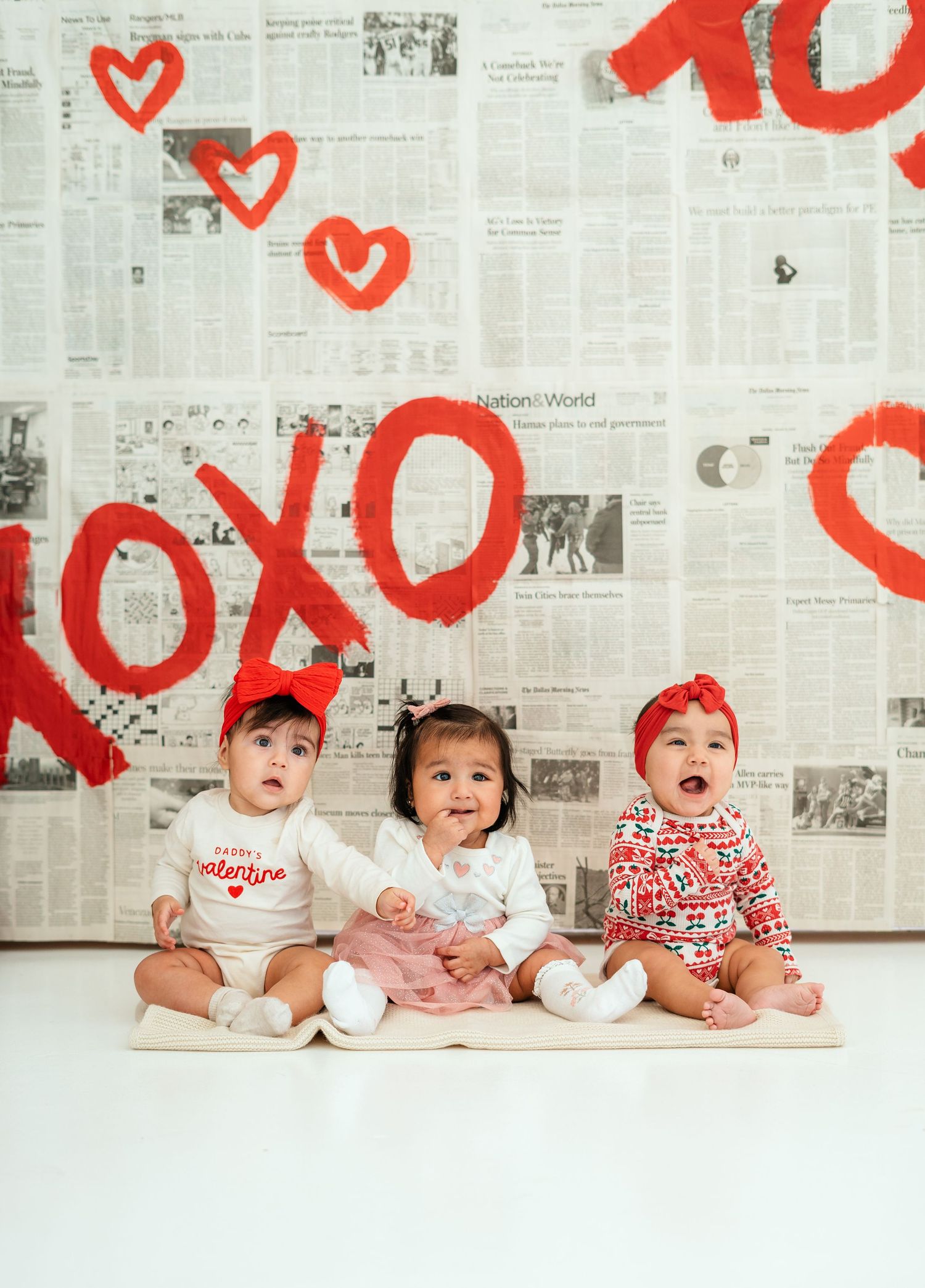 Three cute babies in Valentine's Day outfits posed before a newspaper and hearts backdrop.