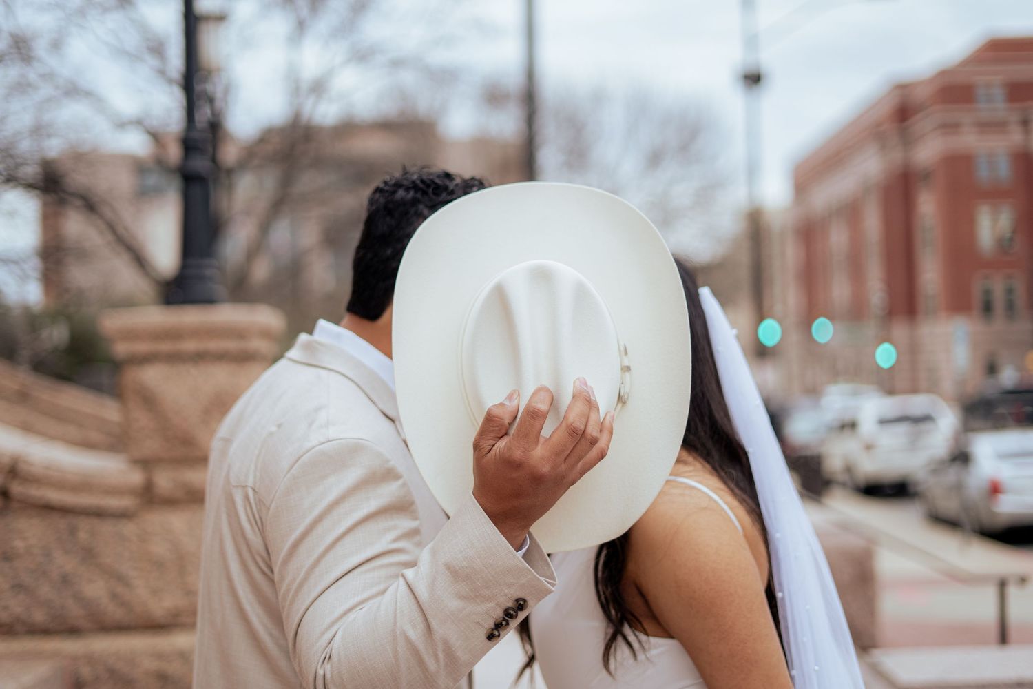 Bride and groom sharing a kiss hidden behind a white cowboy hat on their wedding day outdoors.