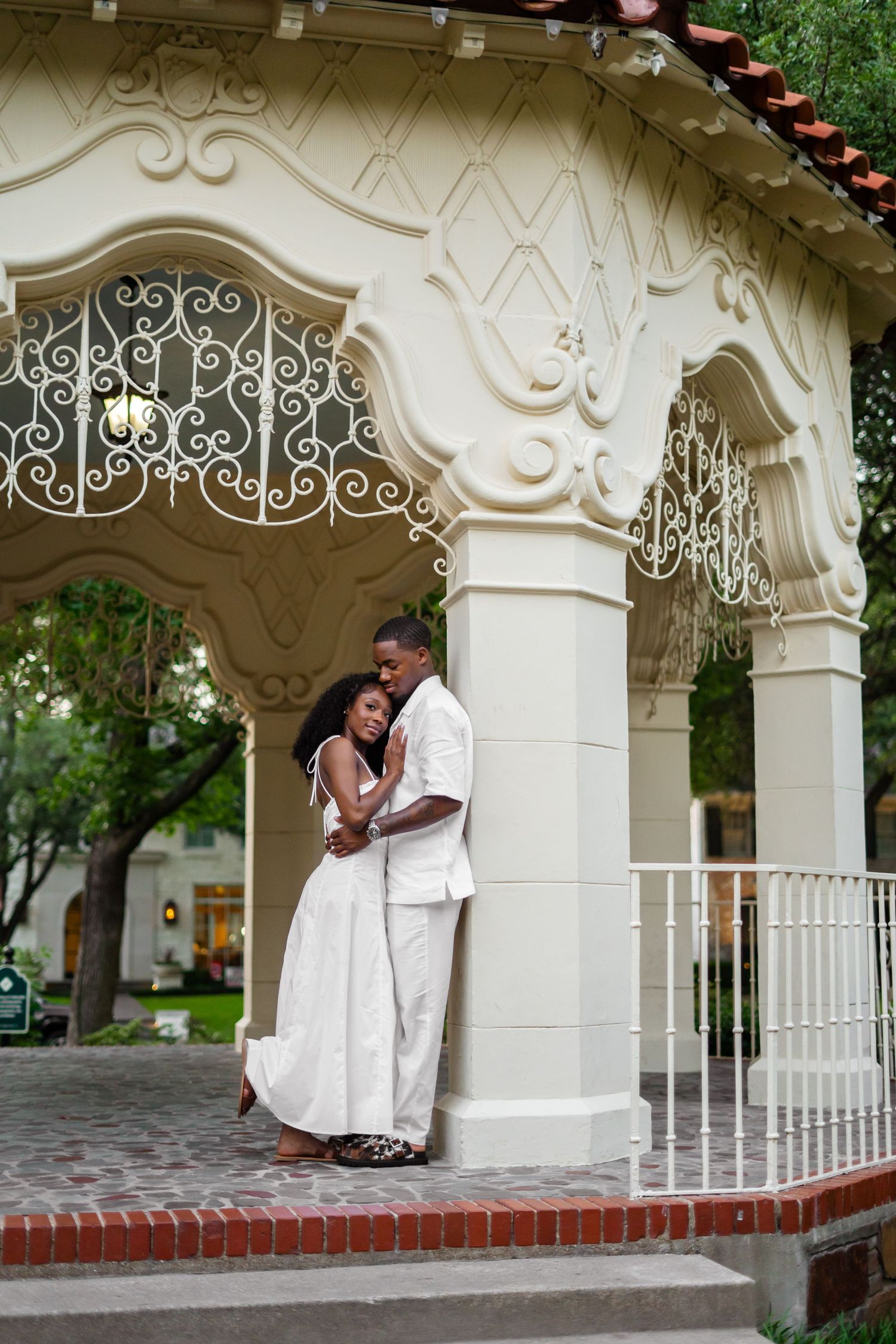 A Black couple in white wedding attire share a tender embrace under an ornate cream-colored pavilion.