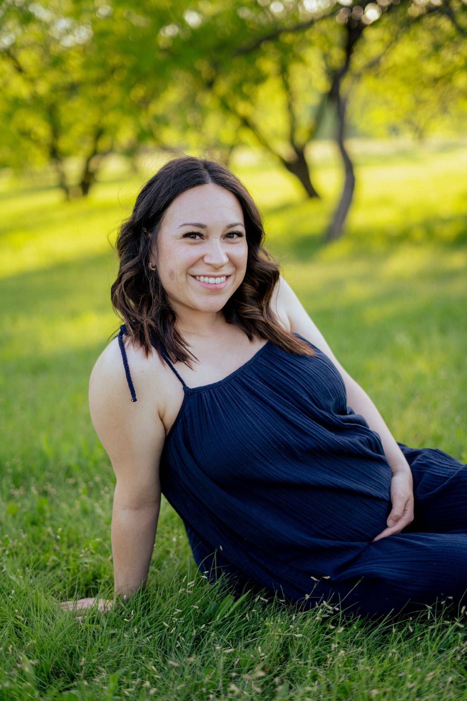 Happy pregnant woman in navy jumpsuit sitting on grass smiling in a sunny park.