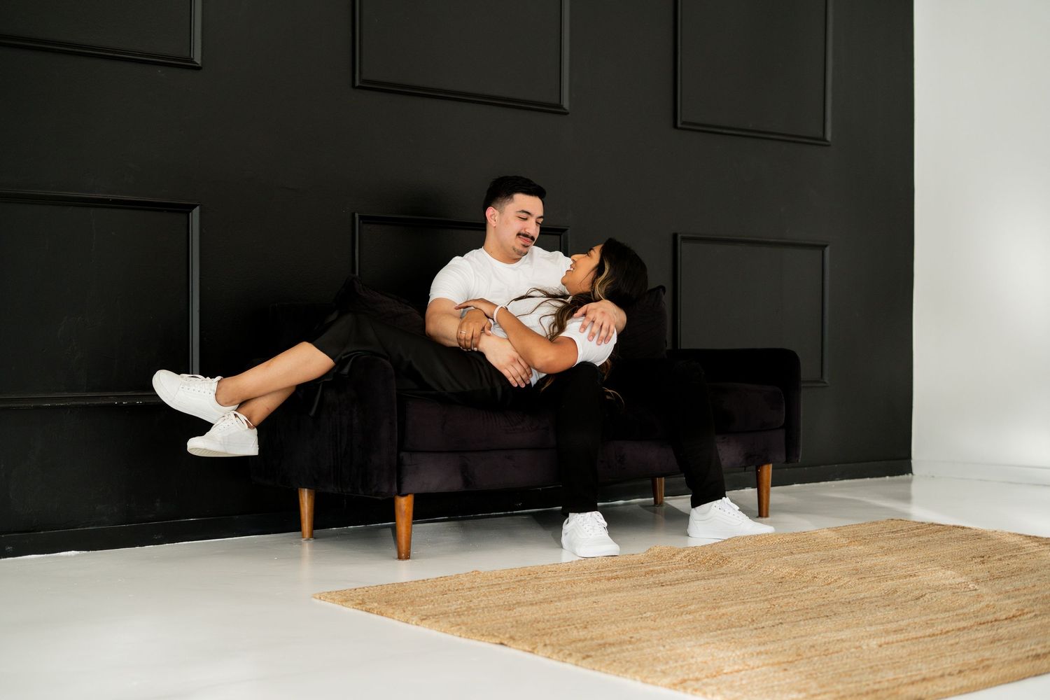 Laughing couple playfully posing together on a black sofa in a modern studio.