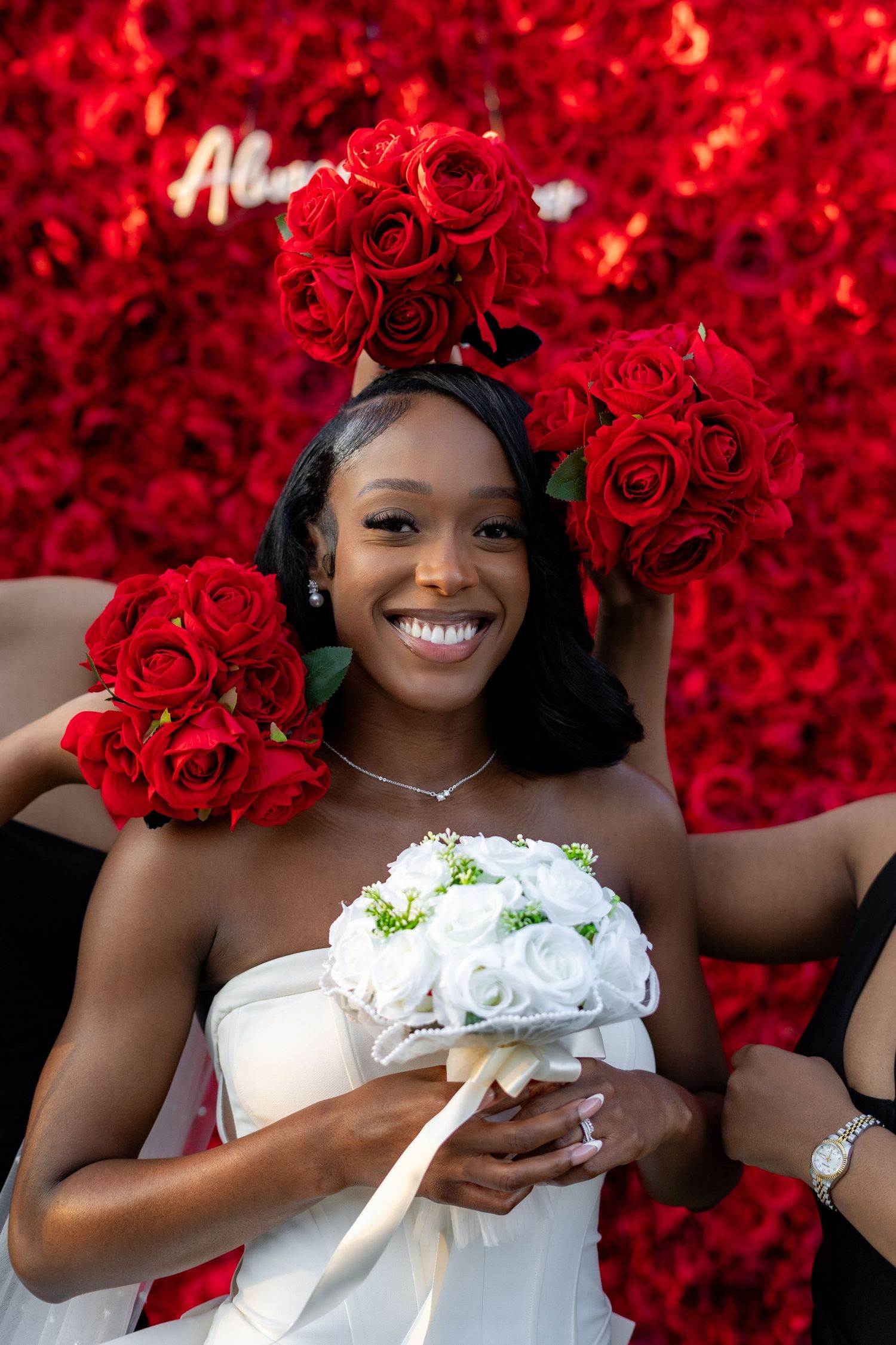 Smiling bride holds white bouquet surrounded by red rose bouquets against a red floral backdrop.