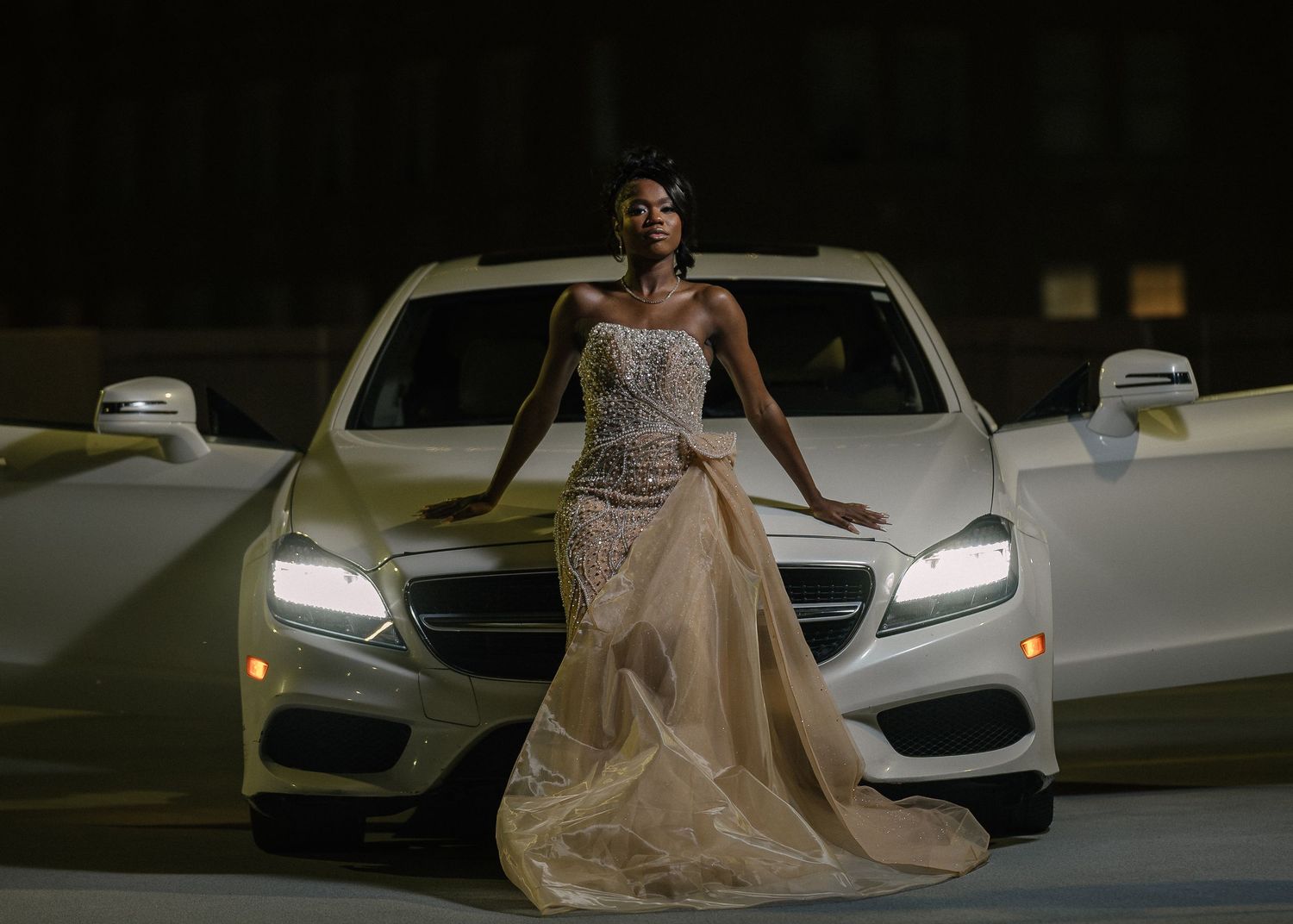 A person in an elegant champagne-colored ball gown poses in front of a white luxury car at night.