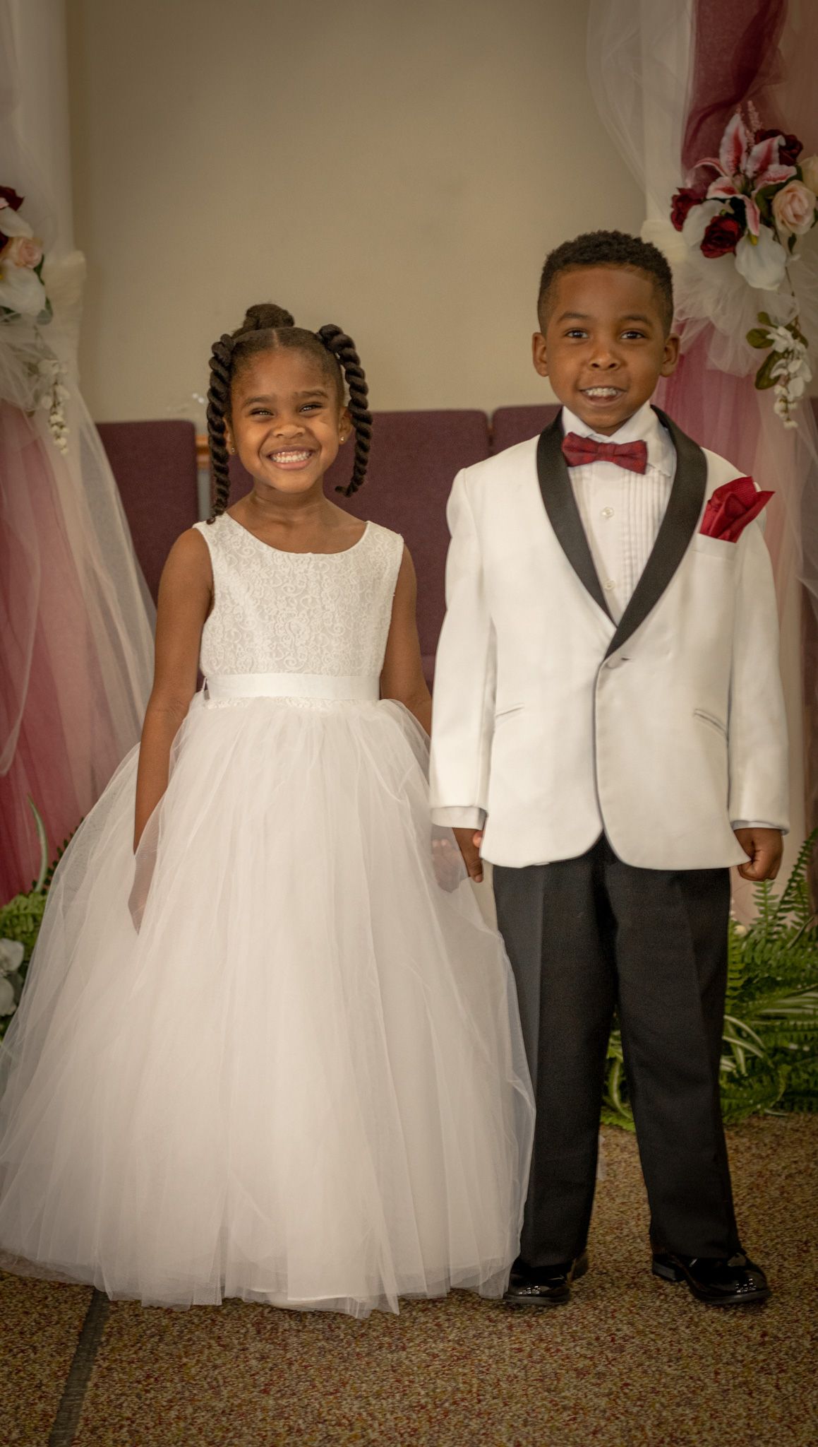 Children in formal white dress and suit attire posing together at wedding event.