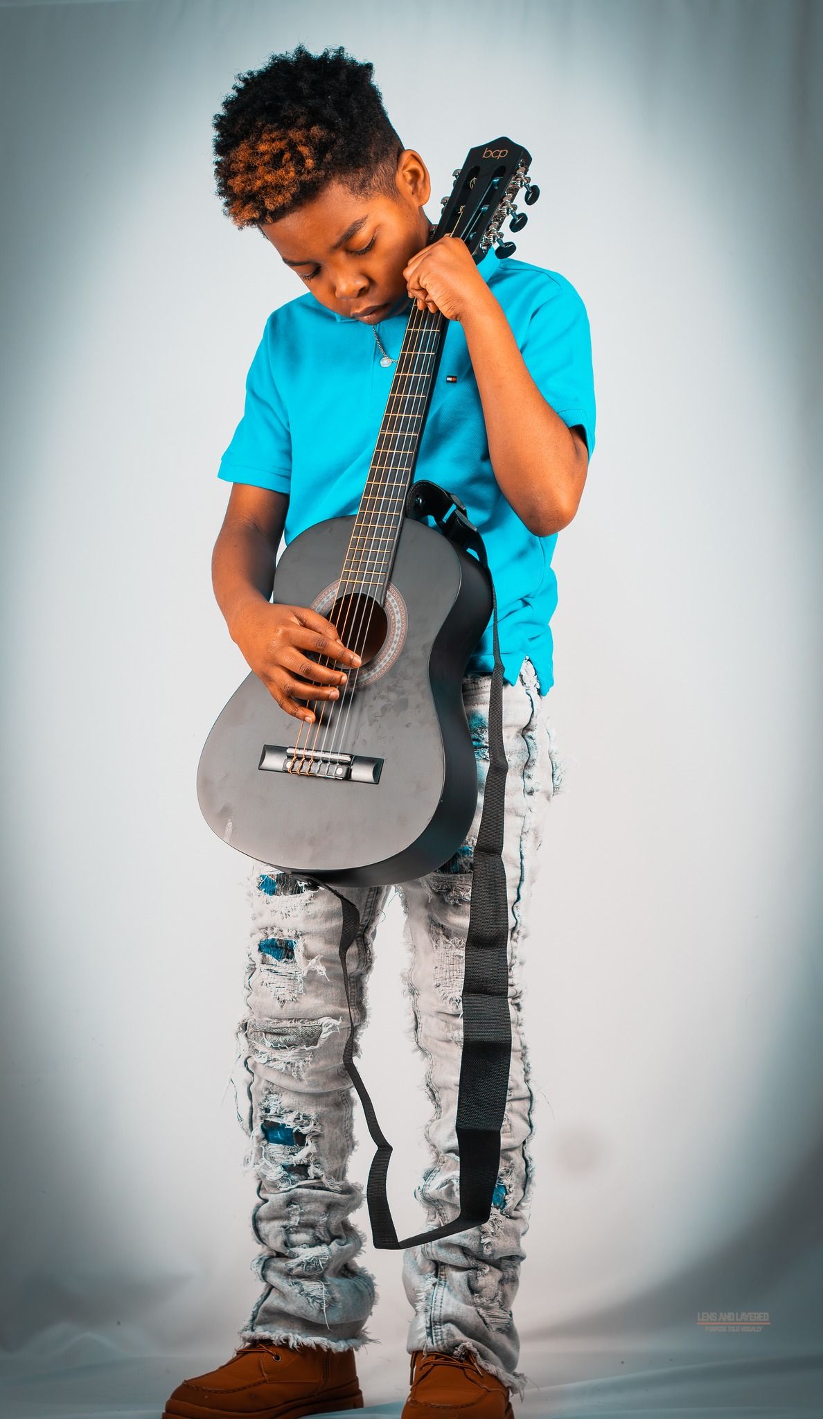 Child in blue shirt holds and plays with a small acoustic guitar against white background.