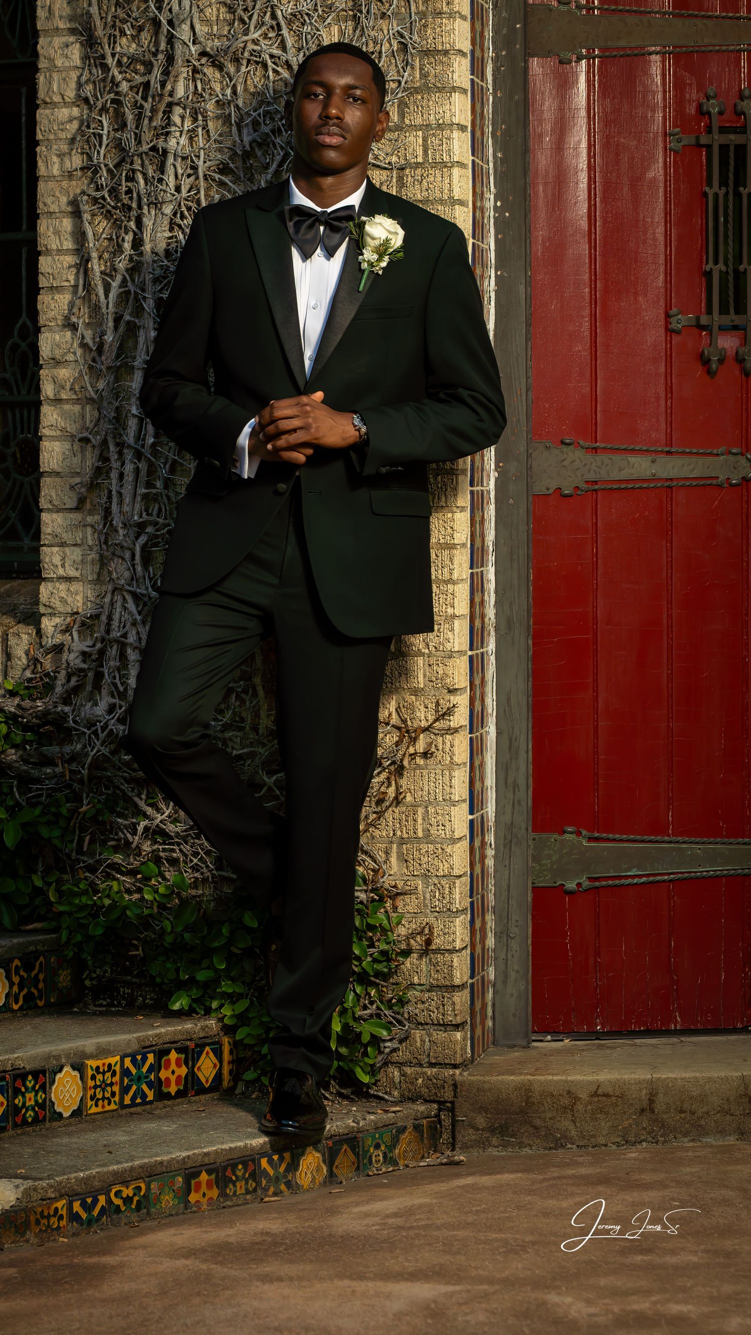 Formal portrait session against stone wall with red door featuring black tuxedo and elegant pose.