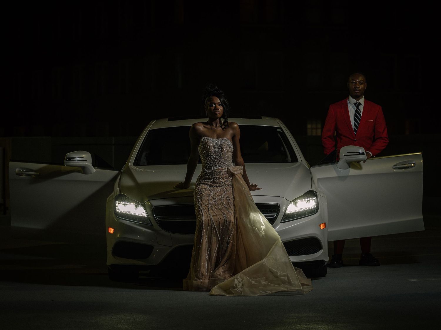 A person in formal attire poses next to a white car at night with dramatic lighting.