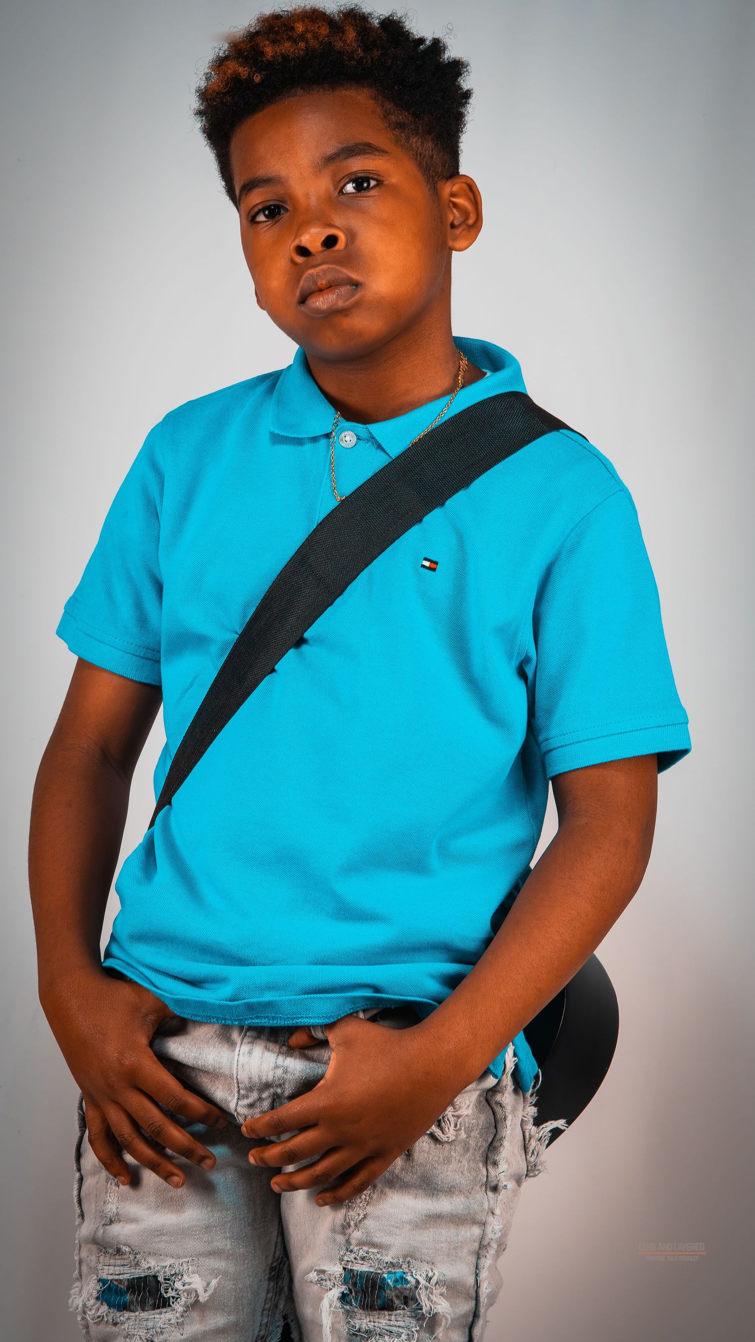 Young person in bright turquoise polo and distressed jeans poses against grey background.