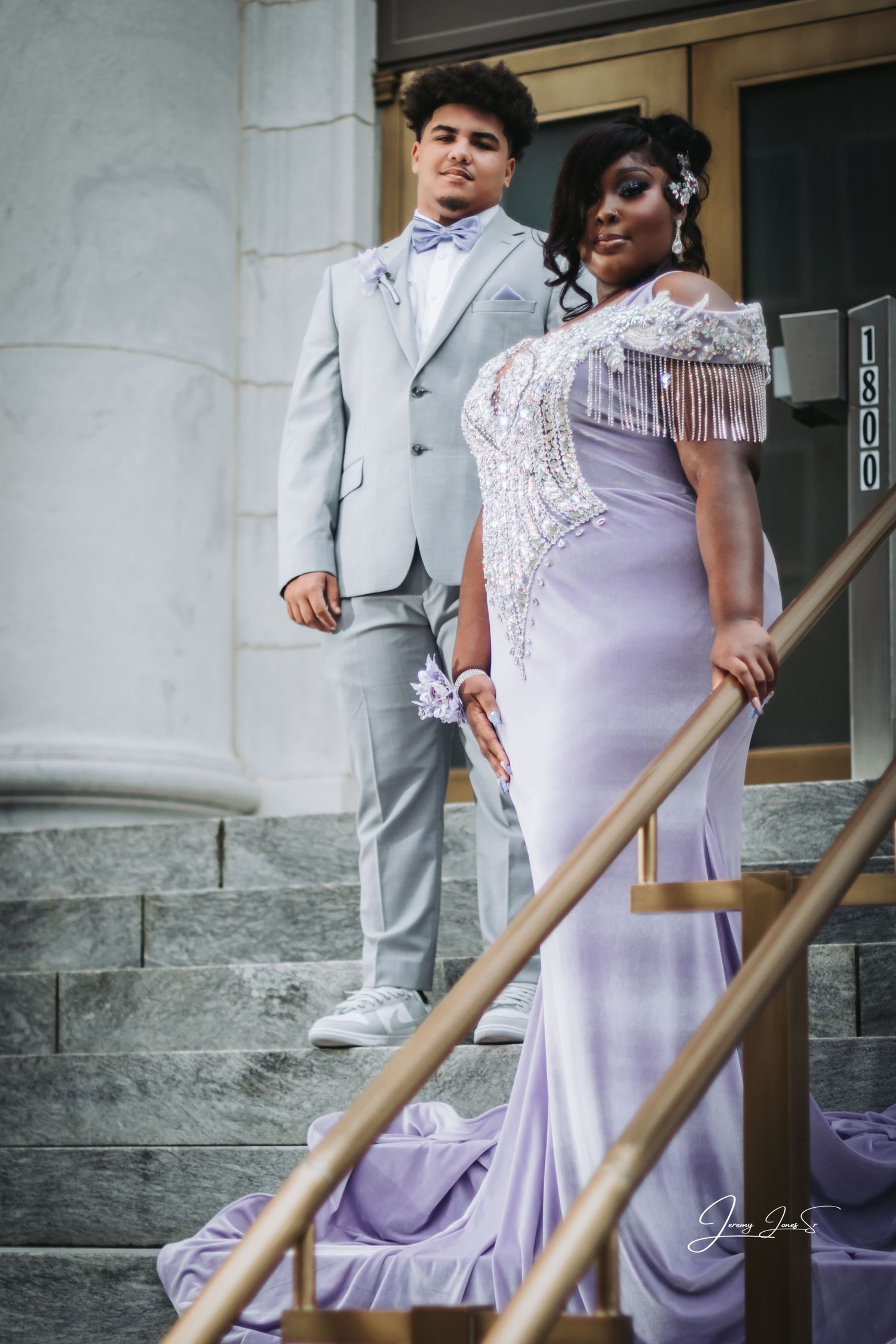 Couple in formal attire pose on stone steps, wearing light gray suit and lavender dress.