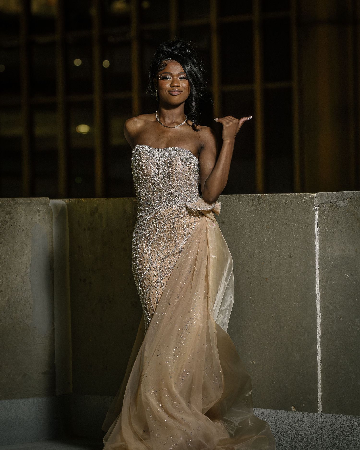 A person poses in an elegant beaded champagne-colored evening gown with a flowing sheer overlay on a balcony at night.
