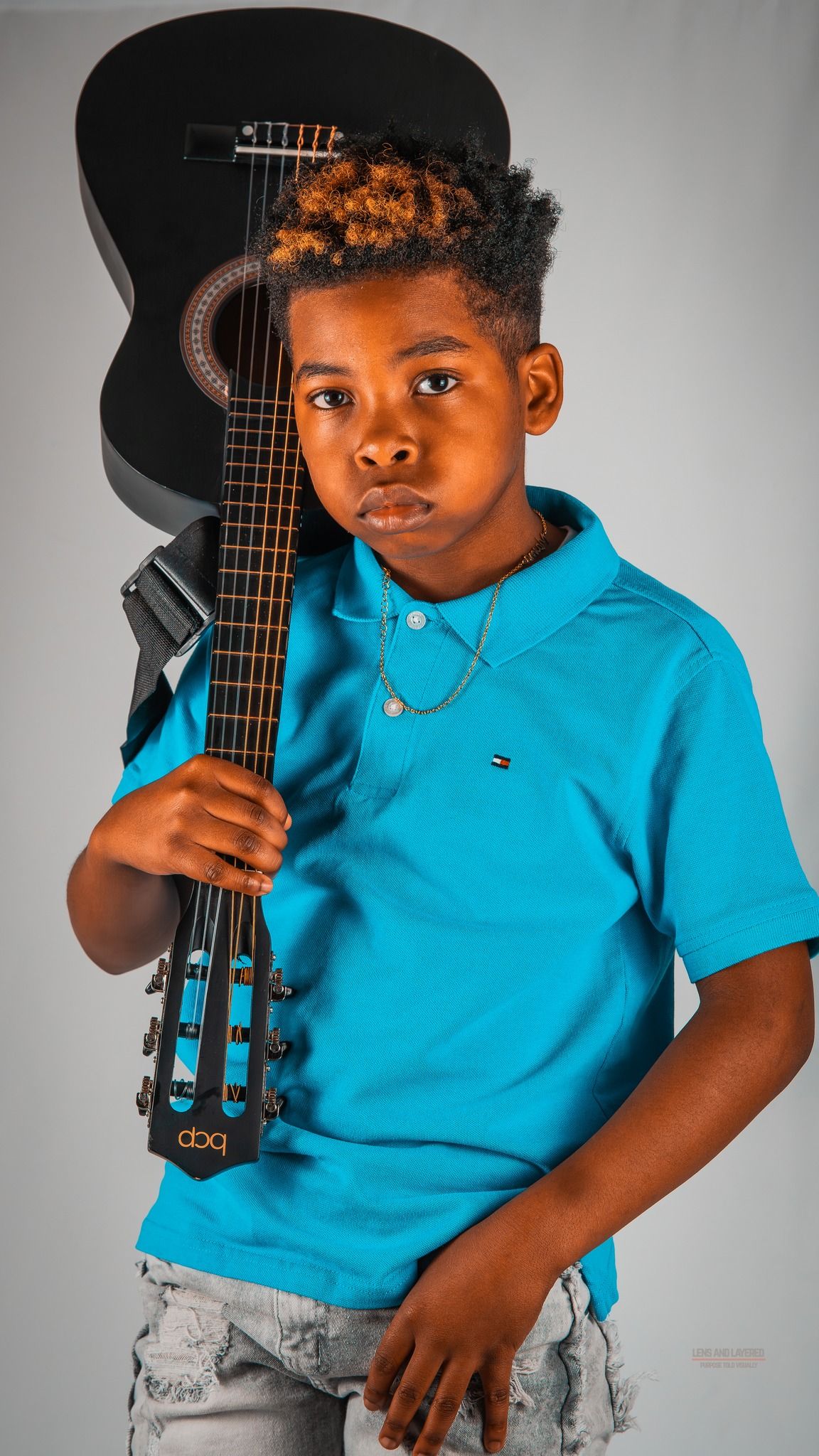 Young musician in bright blue polo shirt poses with black acoustic guitar against gray background.