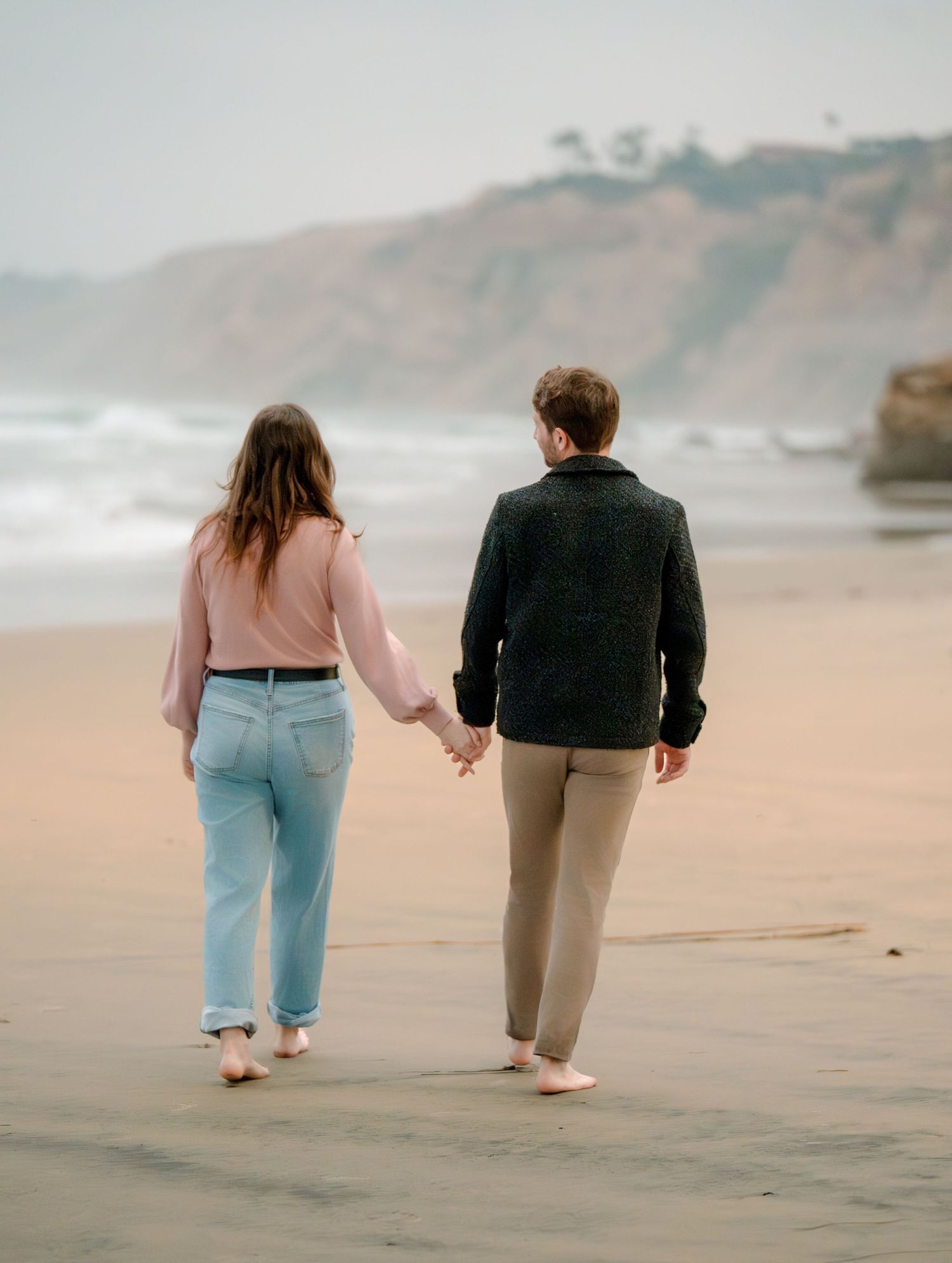 Couple holding hands while walking along scenic coastline during a romantic outdoor engagement photography session.