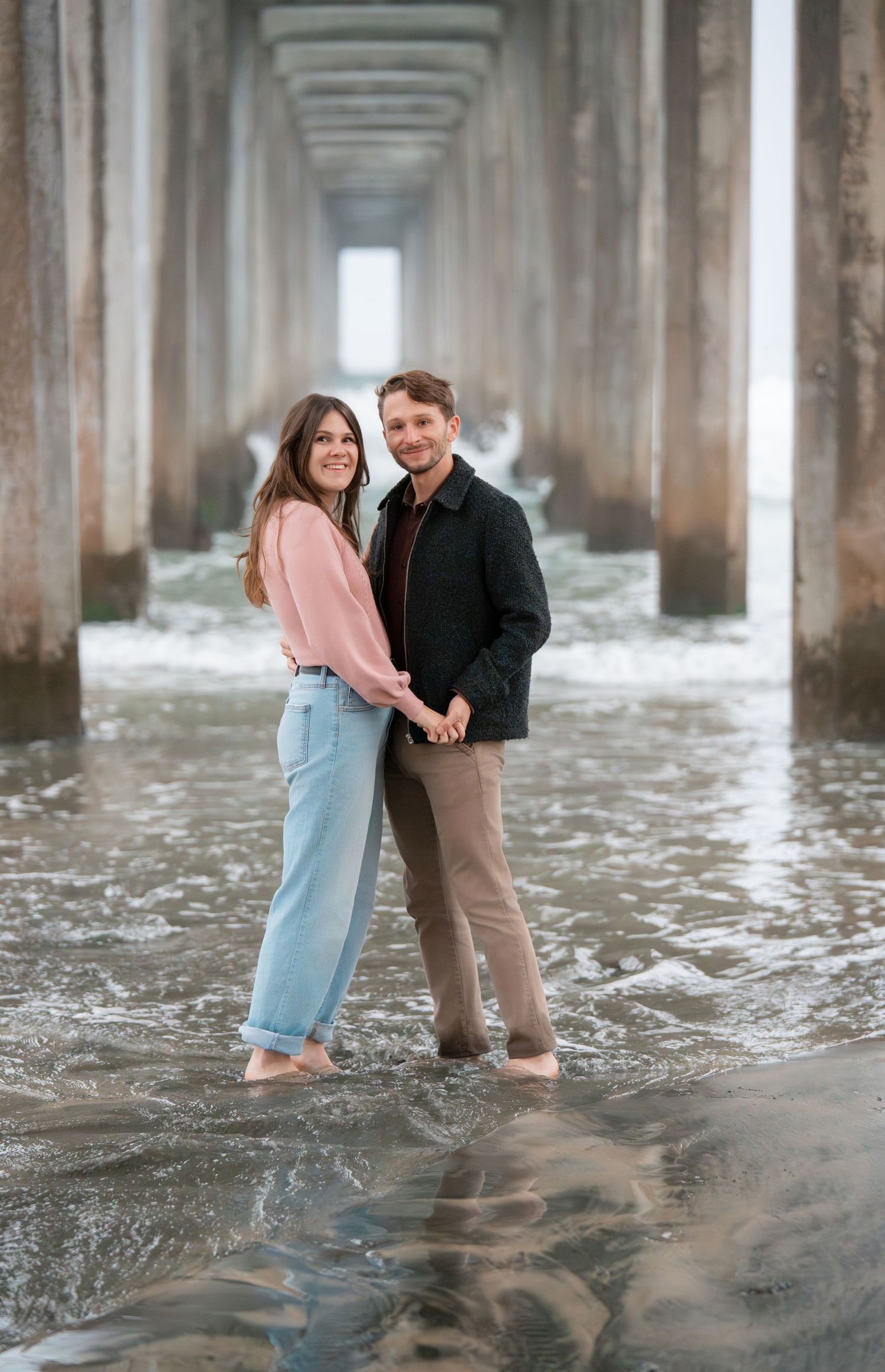 Couple standing under ocean pier during a romantic engagement photography session captured by a couples photographer.