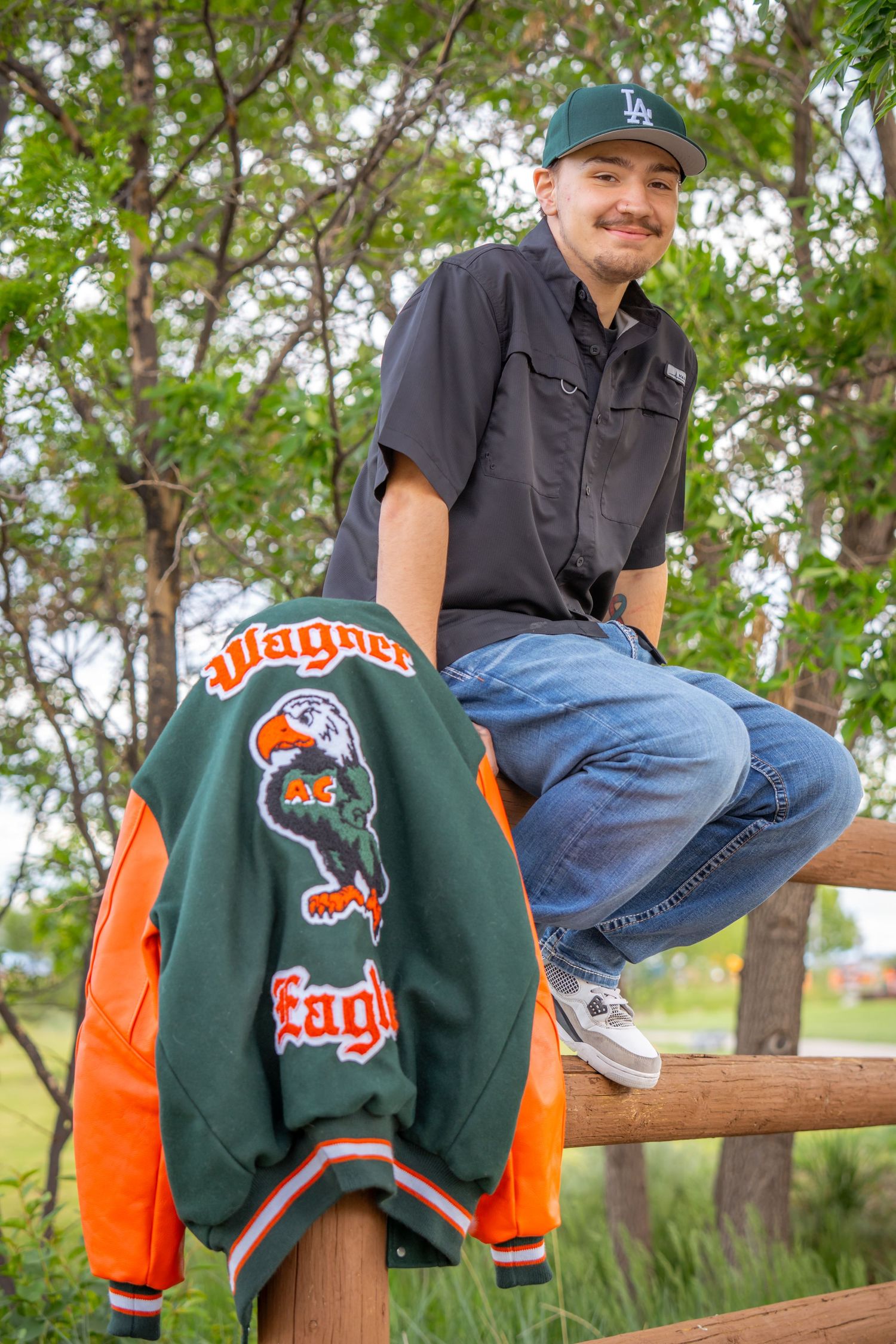 Adams City High School senior portrait sitting on a fence with a letterman jacket during a natural outdoor senior photo session in Colorado.