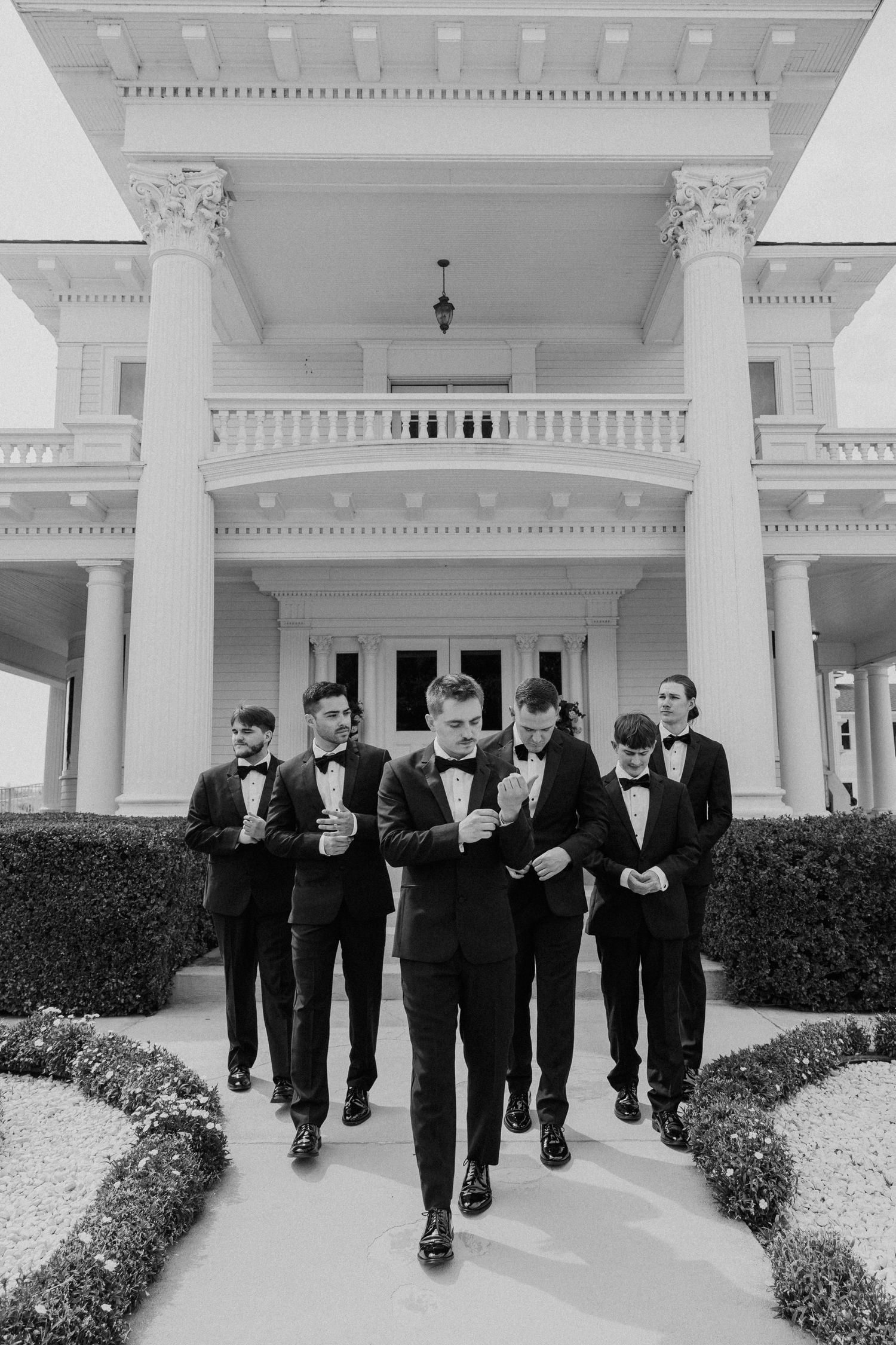Groomsmen in black tuxedos walk together along a white columned walkway at an elegant venue.