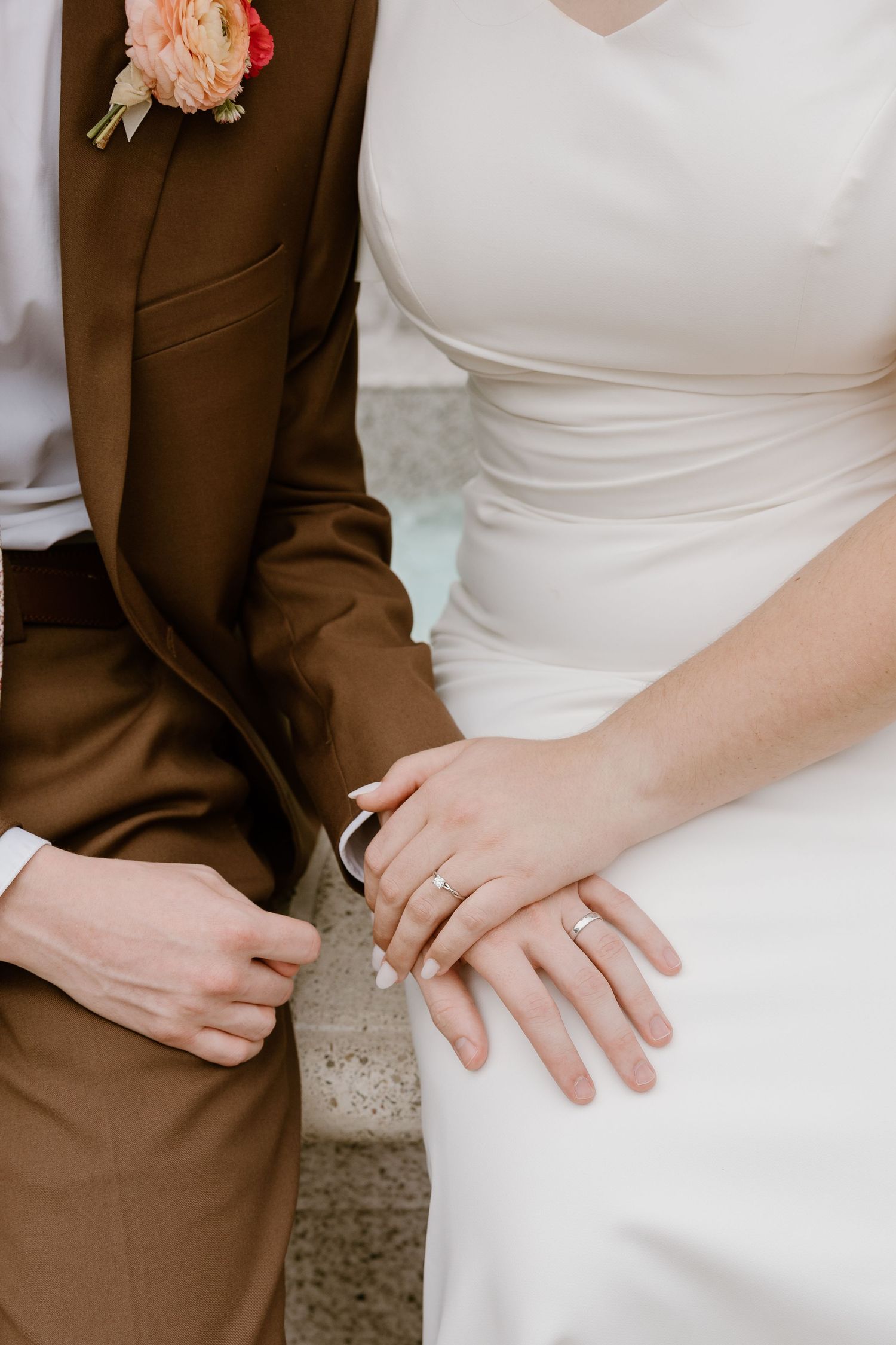 Close-up detail of wedding couple's hands resting together showing white dress and brown suit attire.