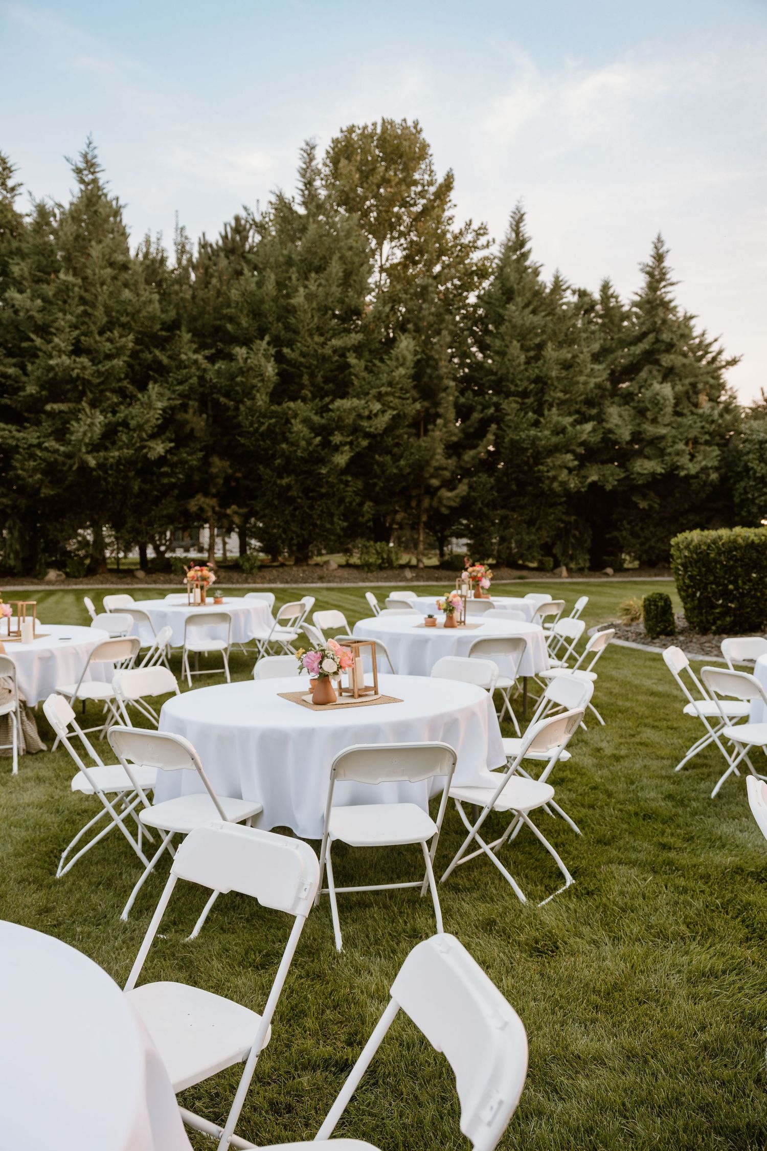 White round tables and folding chairs set up on a lawn for an outdoor wedding reception with evergreen trees in background.
