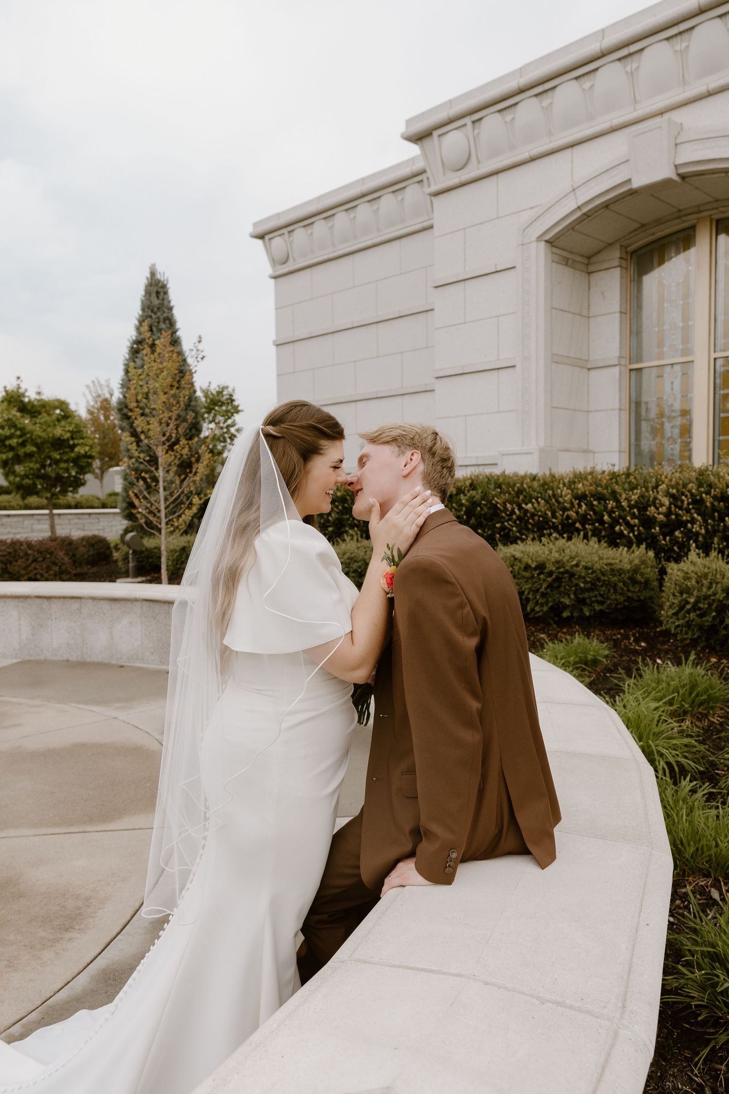 A bride in a white gown and veil shares a tender moment with their partner in a brown suit outside a stone building.