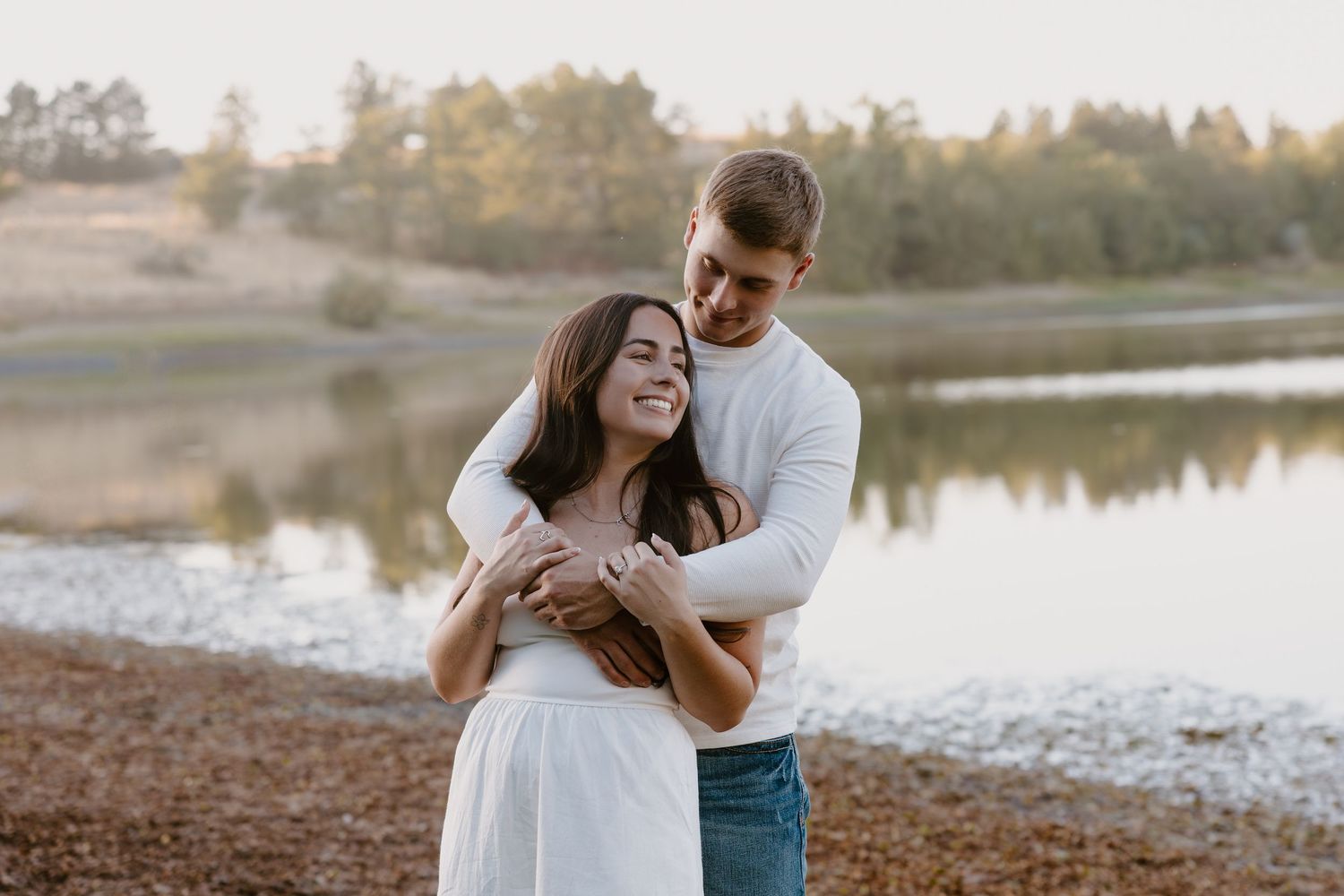 A couple embraces lovingly by a tranquil lake during golden hour sunset.