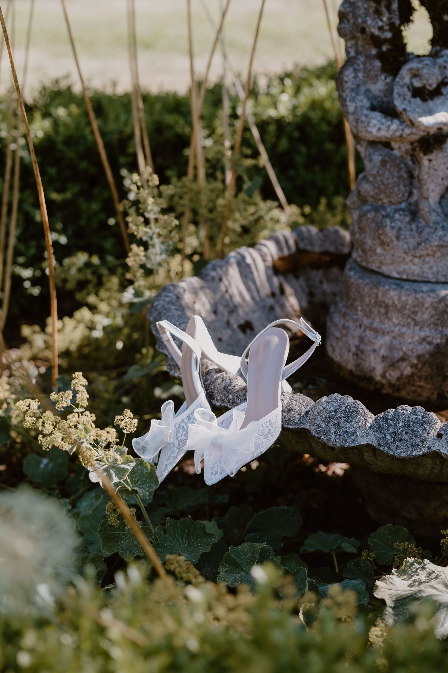 White bridal heels placed among delicate flowers and garden decor with a stone fountain in the background.