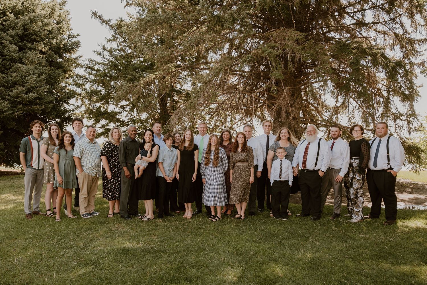 Large family group of people in casual formal attire pose together outdoors on grass with evergreen trees in background.