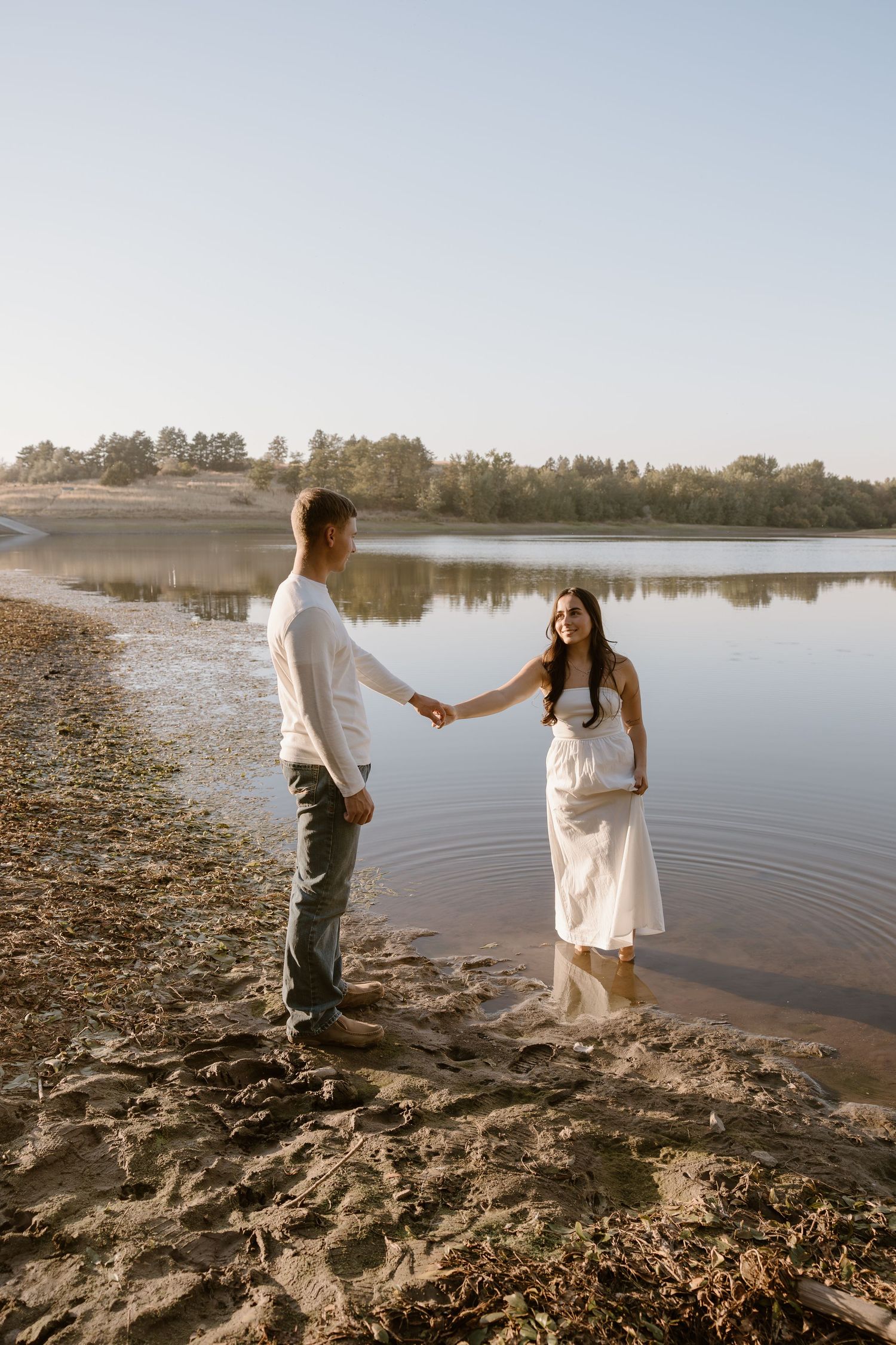A couple holds hands on a serene lakeshore at sunset with calm water and trees in the background.