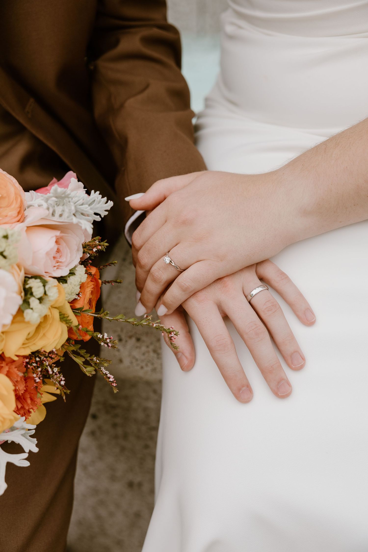 Close-up of wedding couple's hands with rings and peach-colored floral bouquet against brown and white attire.