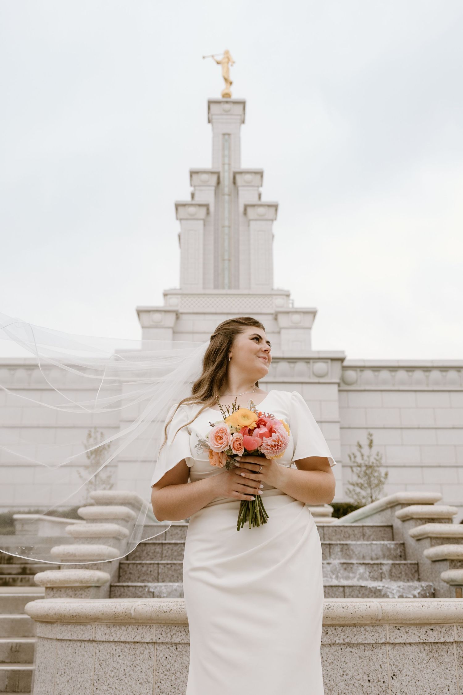 Bride in elegant white dress holds coral bouquet while standing on stone steps of Mormon temple on cloudy day.