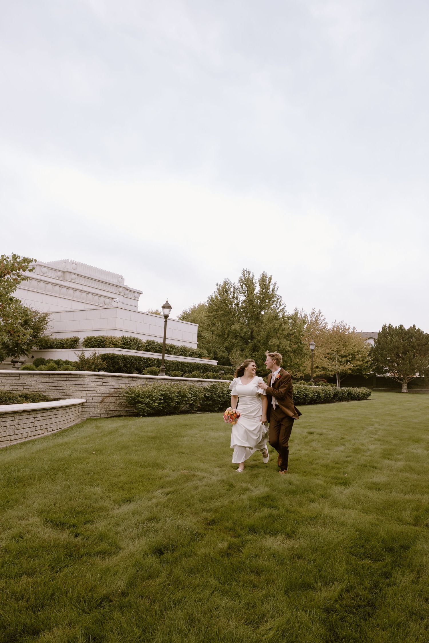 A couple in wedding attire walks across a grassy lawn with a modern architectural building in the background.