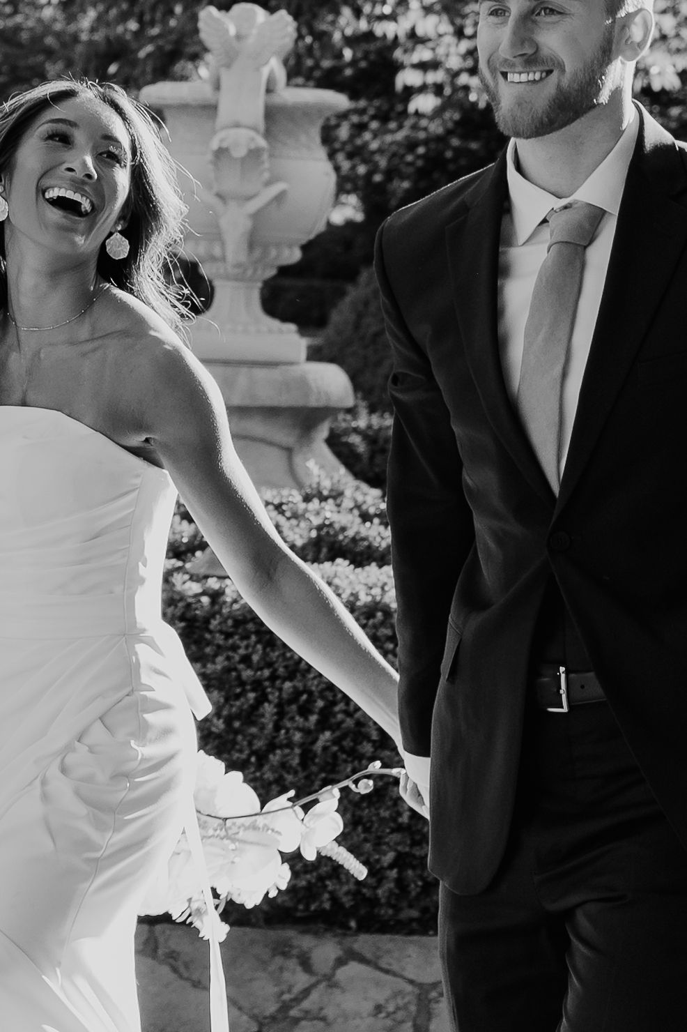 A joyful black and white wedding photo showing a couple laughing together in formal attire near an ornate garden fountain.