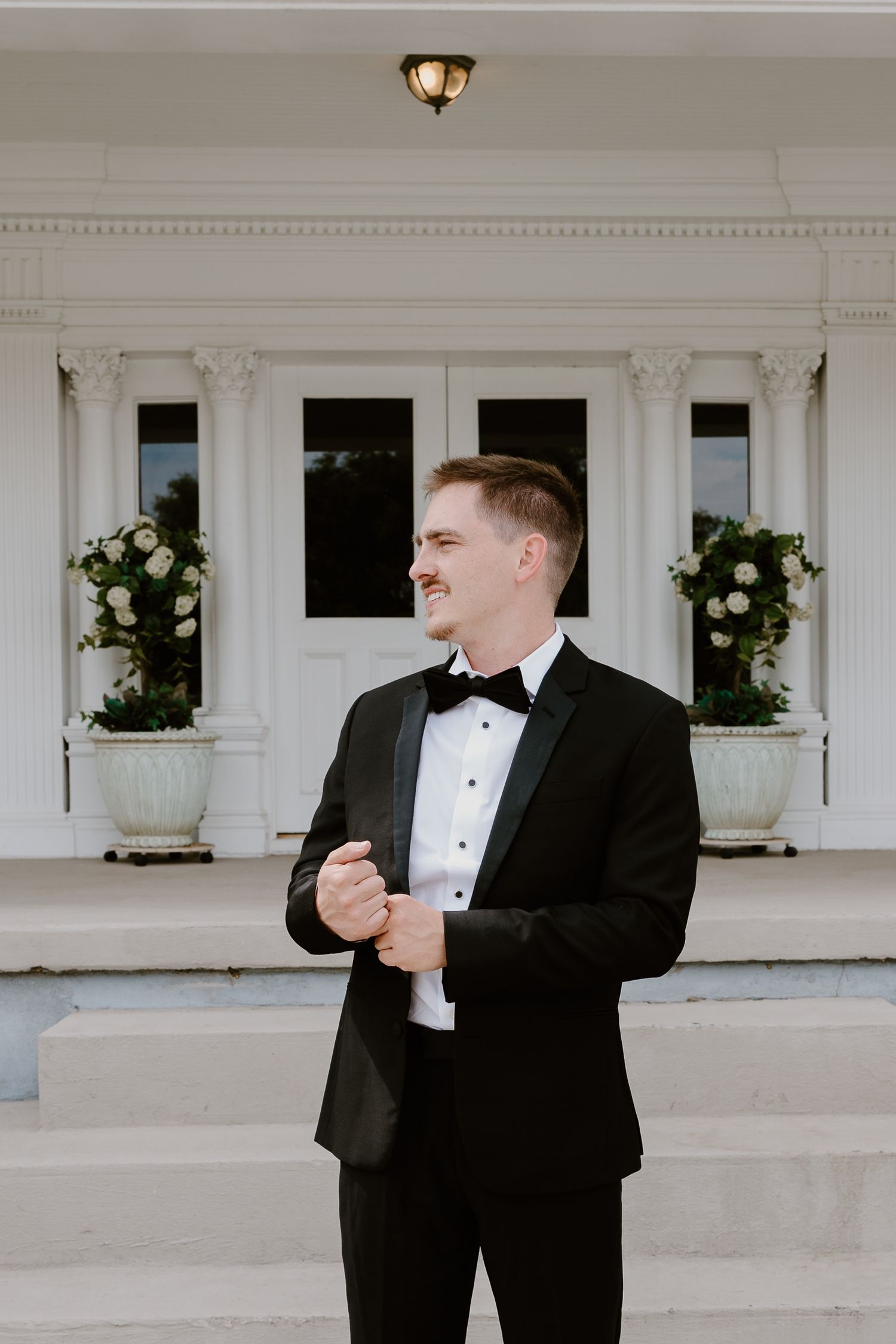 A person in a classic black tuxedo and bow tie smiles while standing in front of a white columned entrance with floral decor.