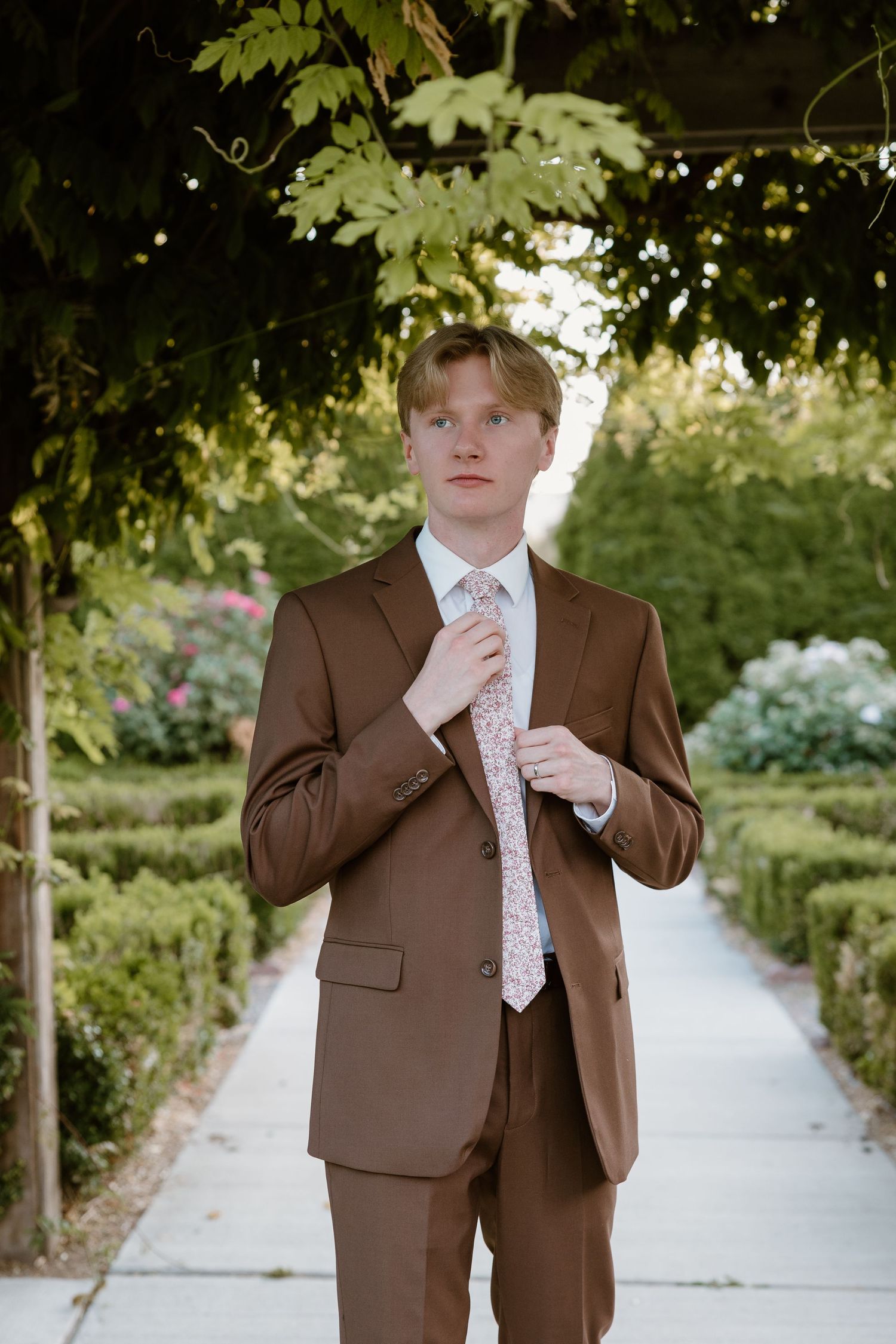 A person in a brown suit and polka dot tie stands in a garden path surrounded by lush greenery and flowers.