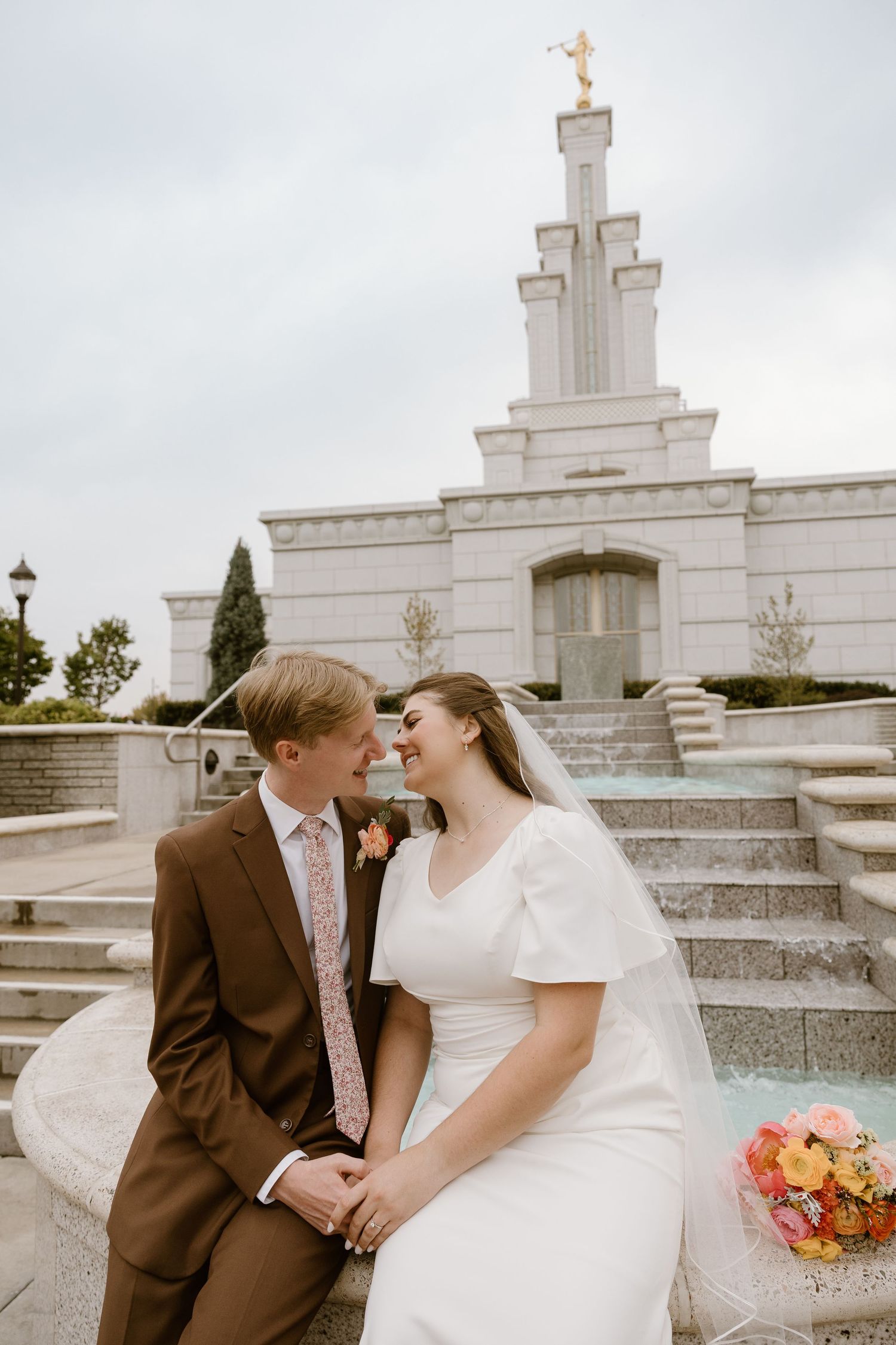 A couple shares a tender moment on stone steps outside a white temple building with a floral bouquet.