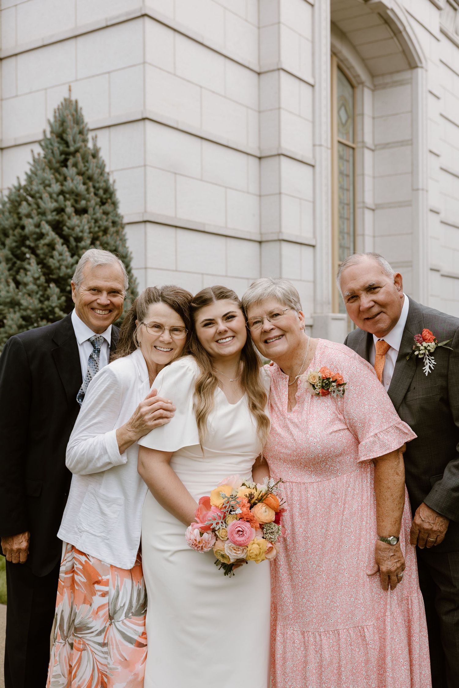 Wedding party members in pink and white attire pose together in front of a white stone building.