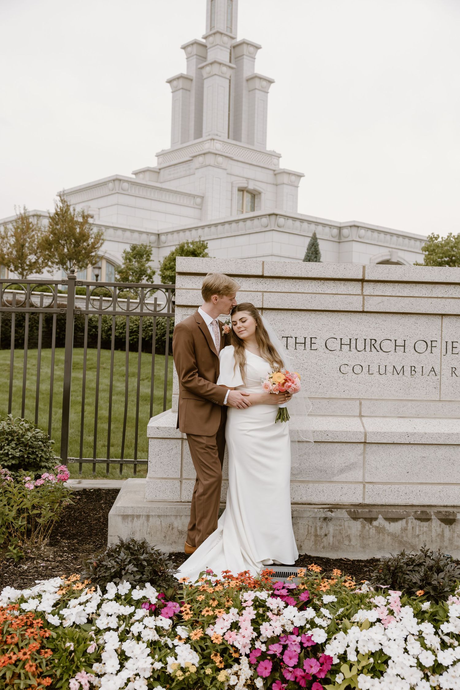 A couple shares a romantic moment in front of a church sign surrounded by colorful flowers on their wedding day.