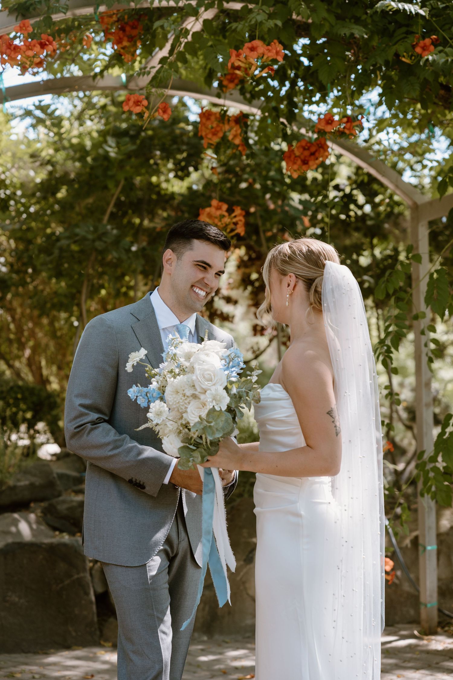 A couple shares joyful moments during their first look on their wedding day beneath trees with red flowers.