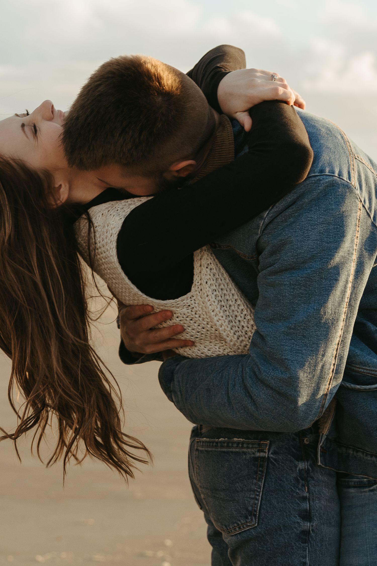 A romantic couple in denim and white sweater embrace passionately against a cloudy sky background on the Oregon Coast beach.