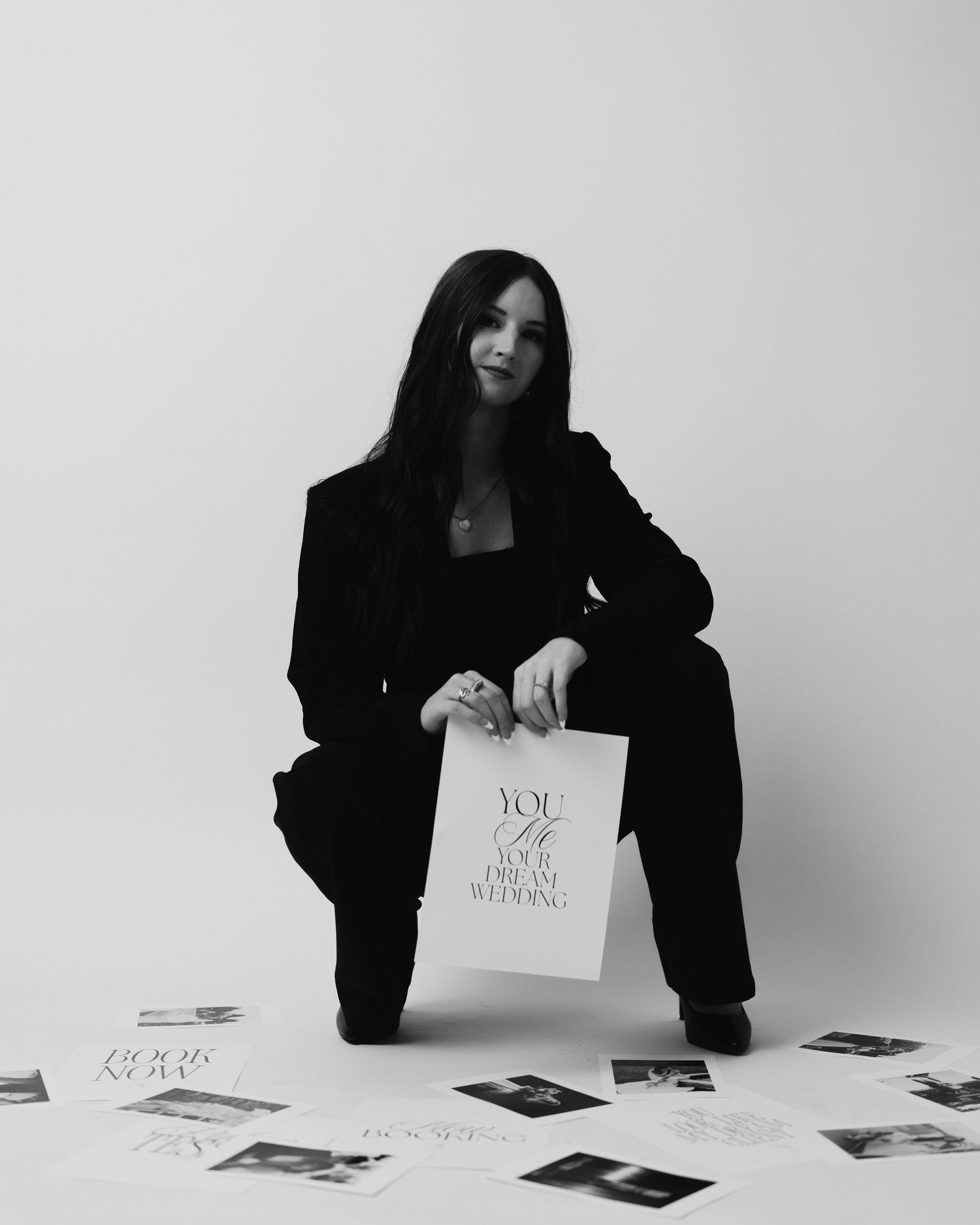A person in black attire squats on the floor holding a sign while surrounded by scattered photographs in monochrome.